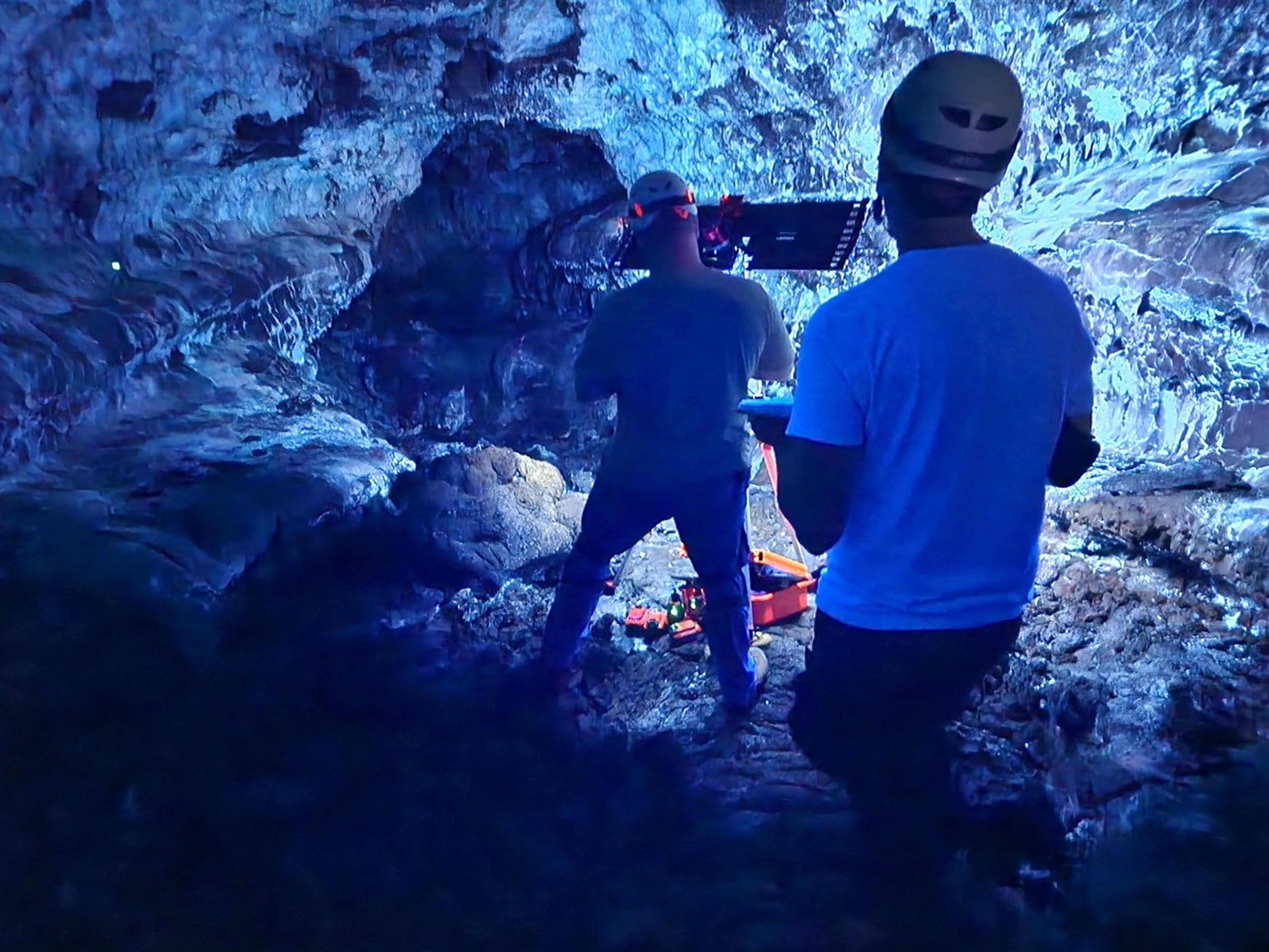 Blue-lit cave interior. Two researchers wearing helmets operate a large, flat light panel mounted on a tripod.