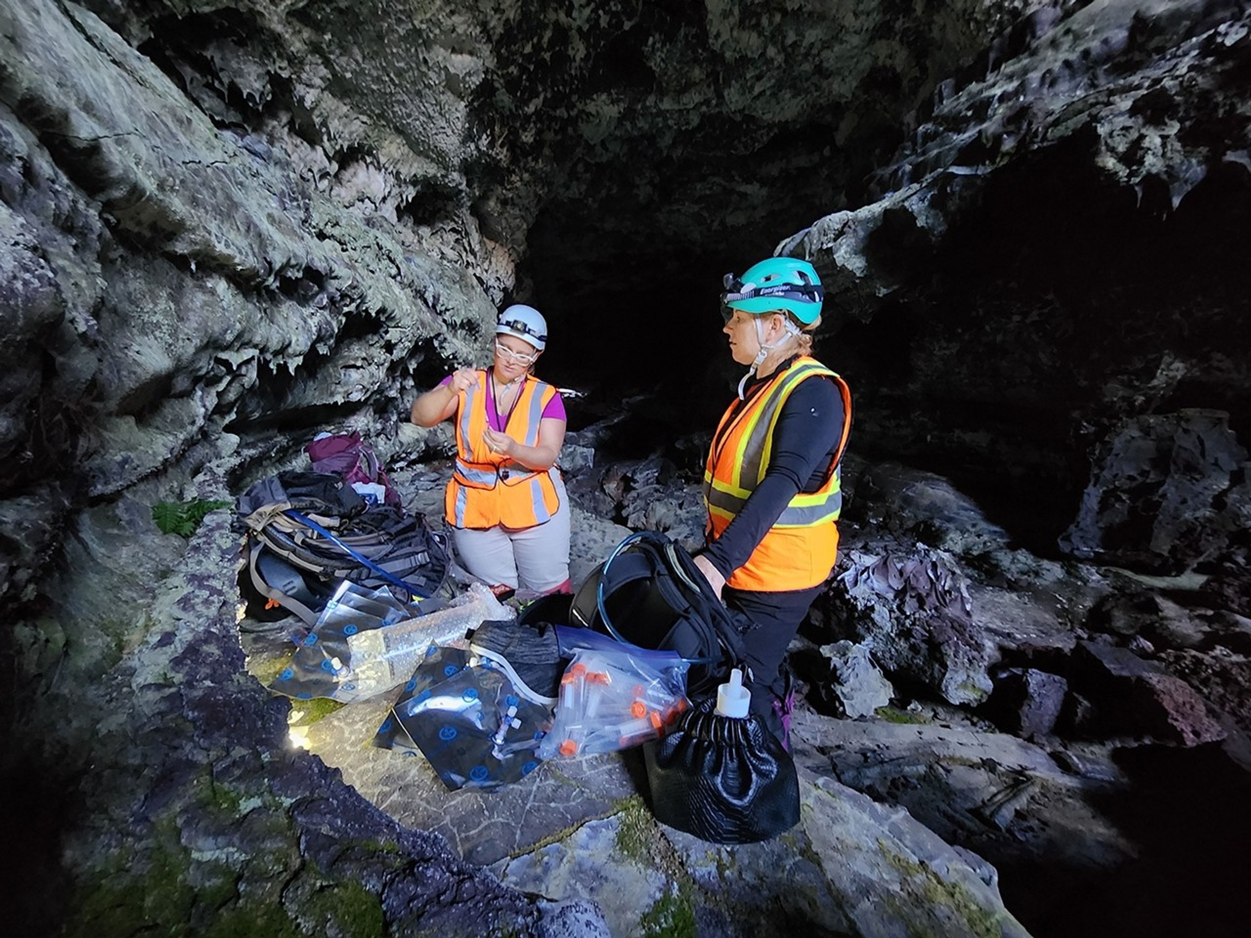 Two women wearing reflective safety vests, helmets, and headlamps in a dark cave. Lab equipment is laid out on the rock shelf in front of them, including small plastic vials, mylar bags, and backpacks.