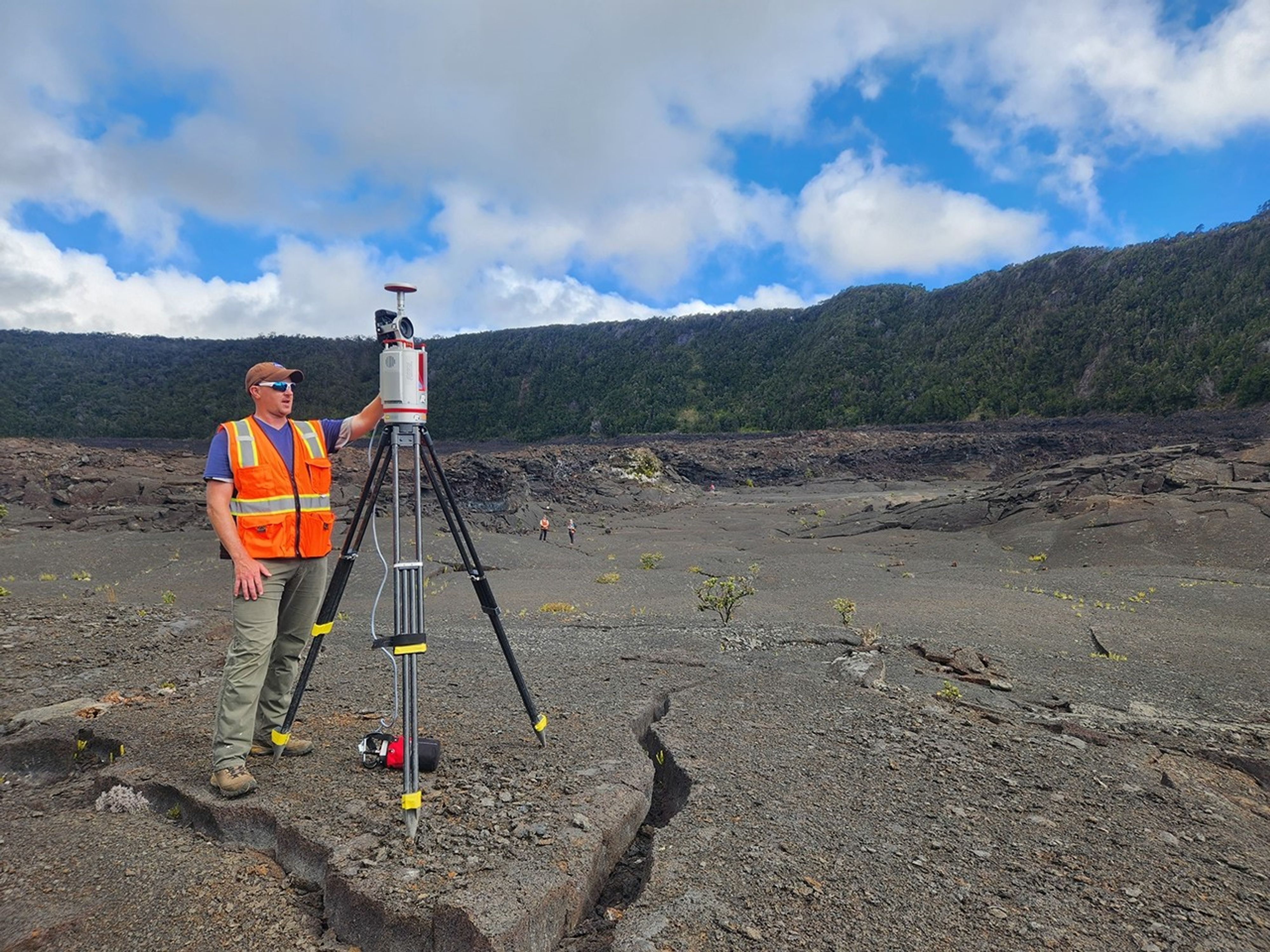 A researcher wearing a high-visibility vest operates a large cylindrical science instrument mounted on a tripod almost as tall as he is, on an expanse of cooled lava with a tree-lined crater rim in the background.