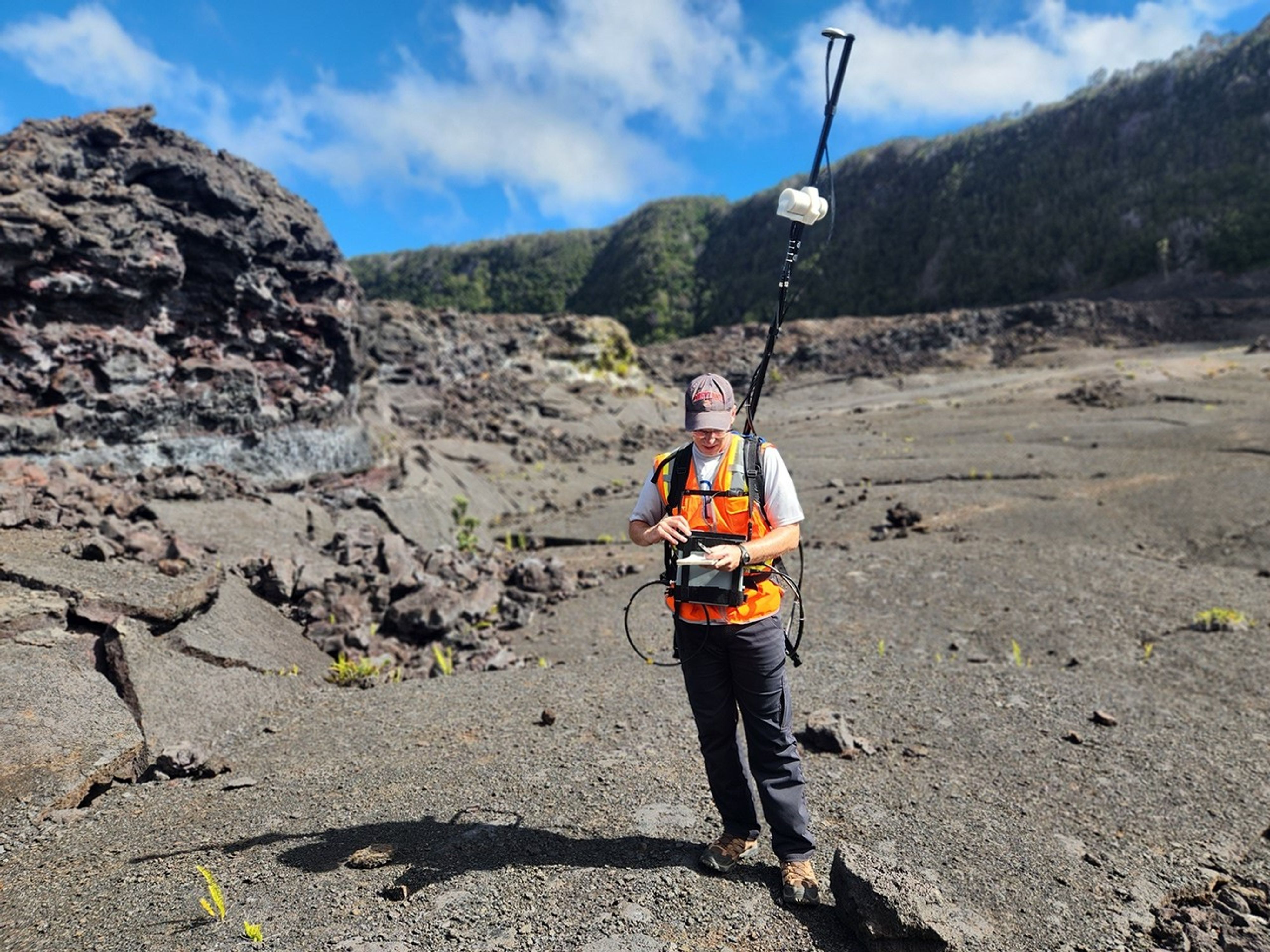 A researcher stands in the bottom of a huge volcanic crater wearing a backpack-mounted science instrument with a long antenna extending several feet overhead.