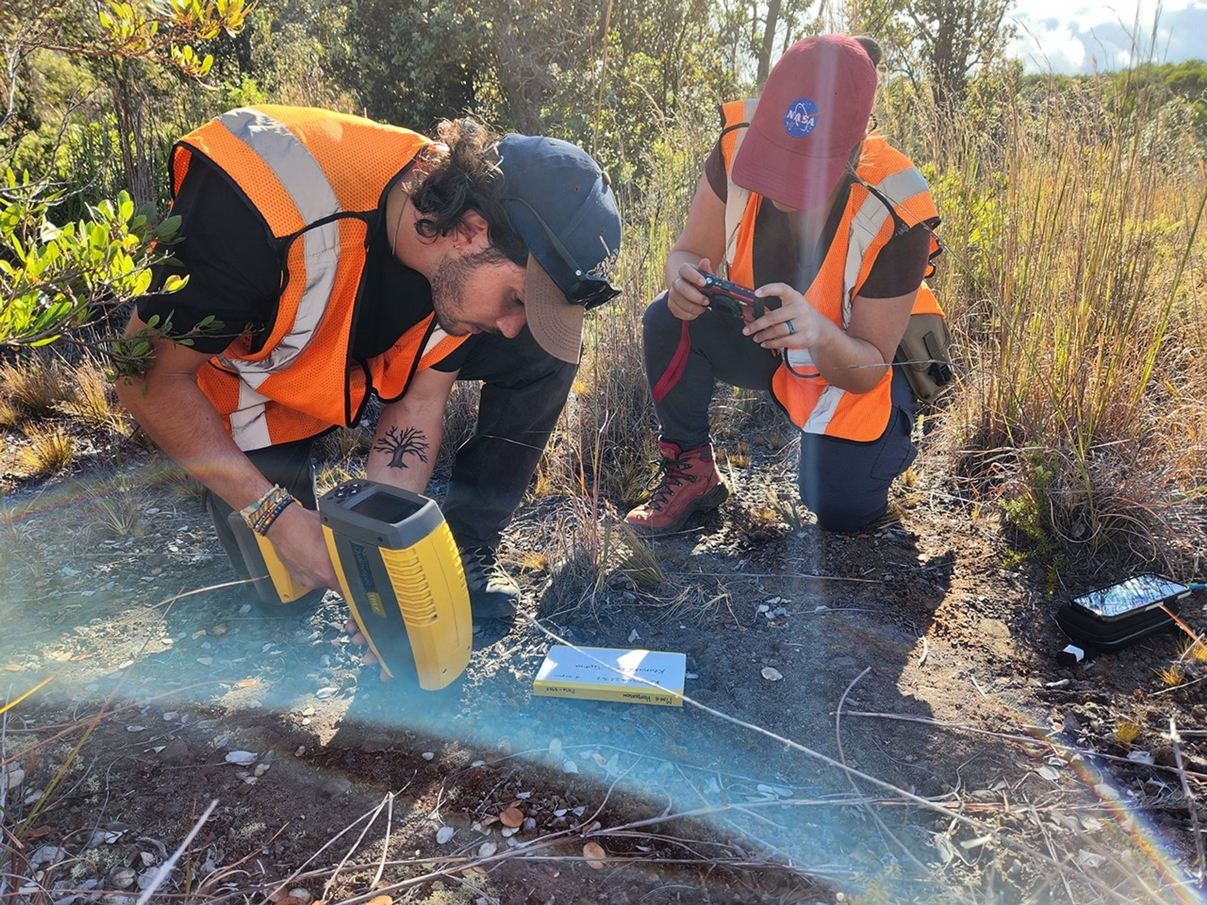 Two researchers wearing safety vests at an outdoor field site. One operates a hand-held device that looks like a yellow blow dryer. The other takes pictures of the process.