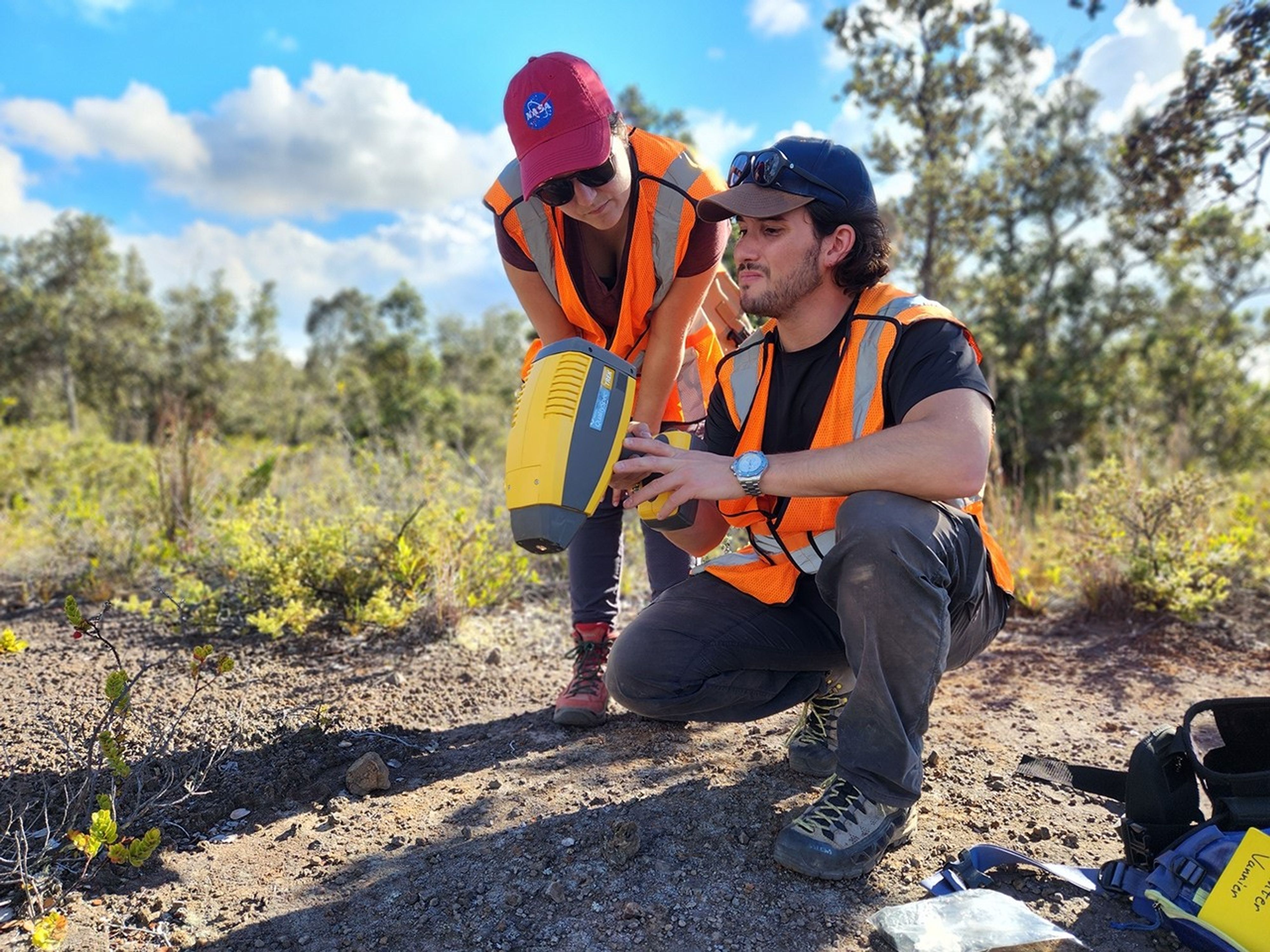 Two researchers wearing safety vests read the output of a hand-held science instrument.