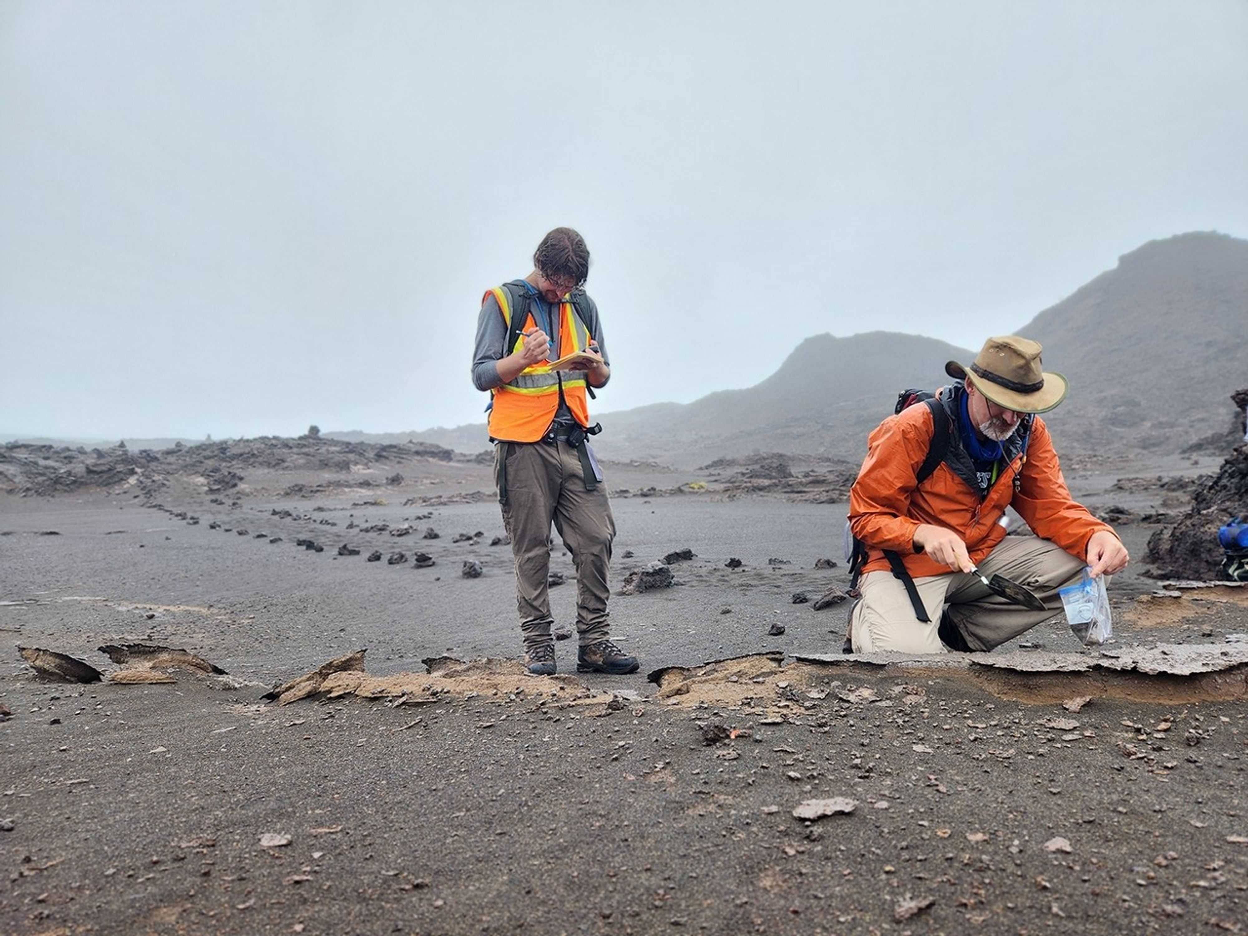 In a barren, rainy landscape, a researcher wearing a bright orange raincoat kneels to scoop a small amount of material from the ground into a plastic bag. His teammate takes notes.