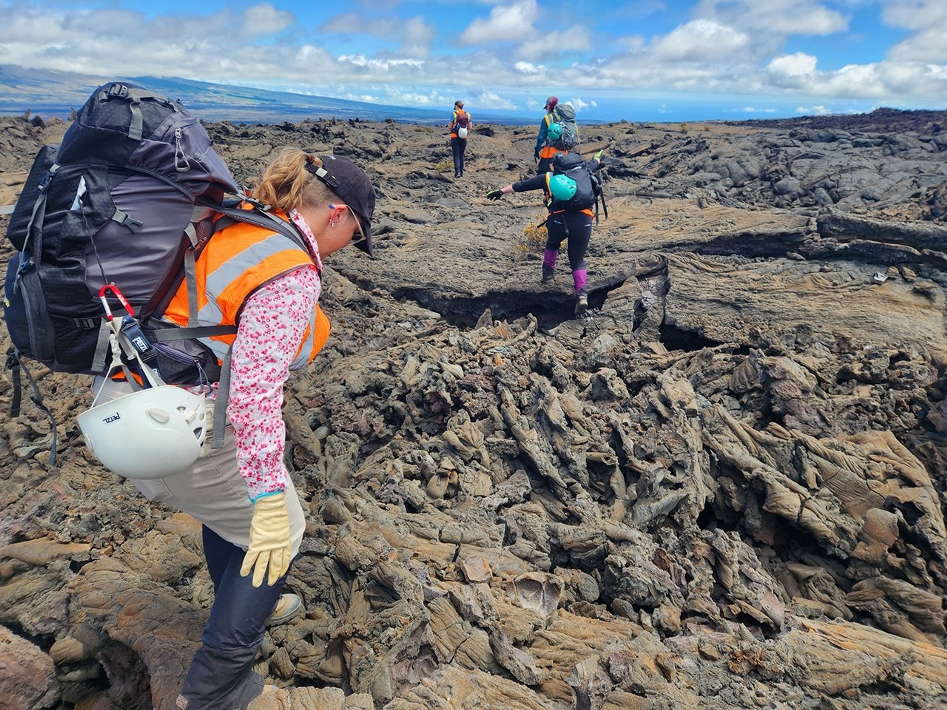 Researchers pick their way across a rugged field of cooled lava. The person in the foreground wears a flowered pink work shirt, a high visibility vest, work gloves, a sun hat, and a large, full backpack. Ahead of her, a teammate steps across a gap in the rocky surface, and more colleagues hike on into the distance.