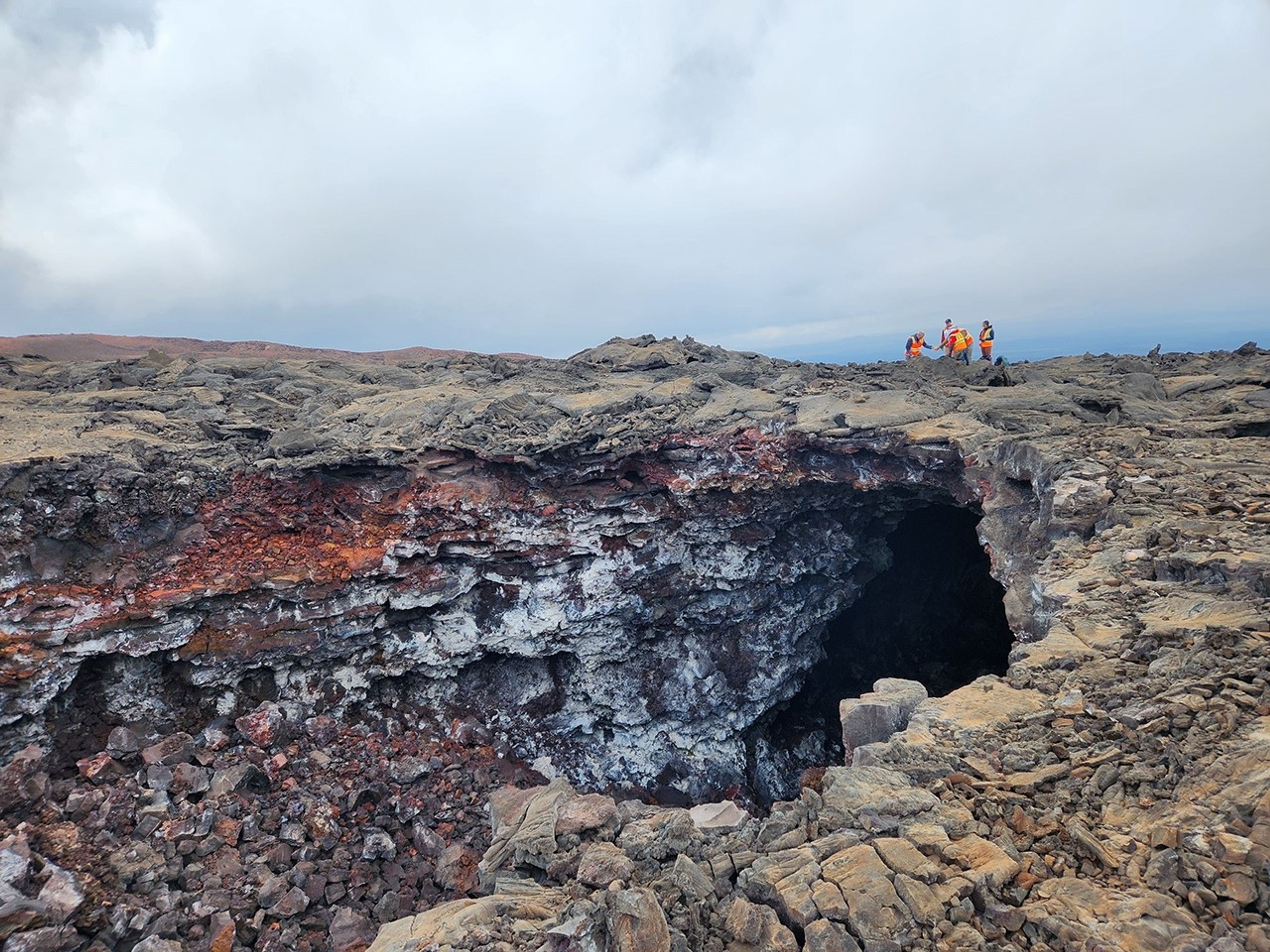 A large, dark cave entrance beneath the surface of a rocky landscape. In the distance, above the cave roof, are several people wearing safety vests.