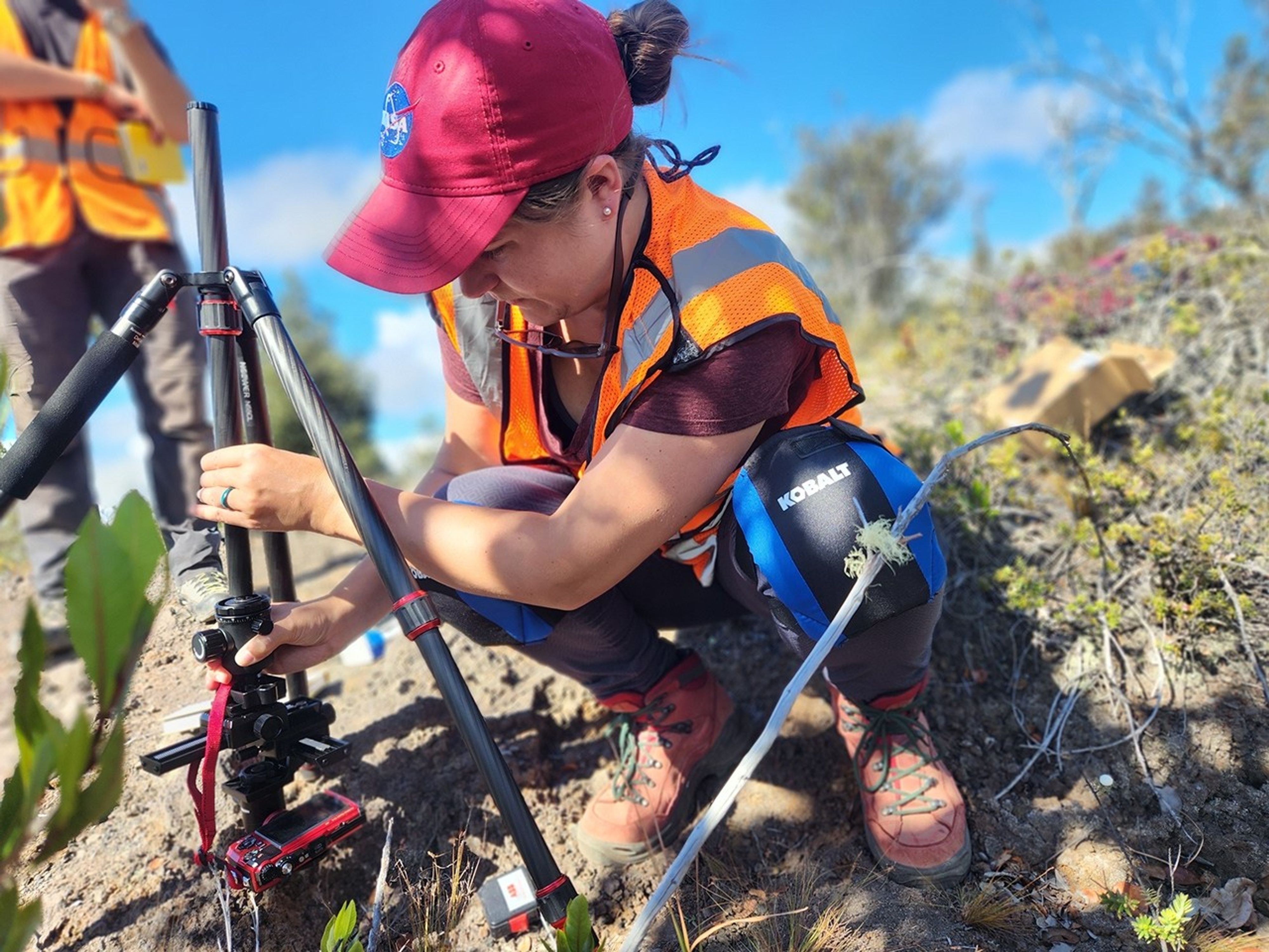 Researcher, wearing a safety vest and knee pads, adjusting an inverted tripod with a camera attached.