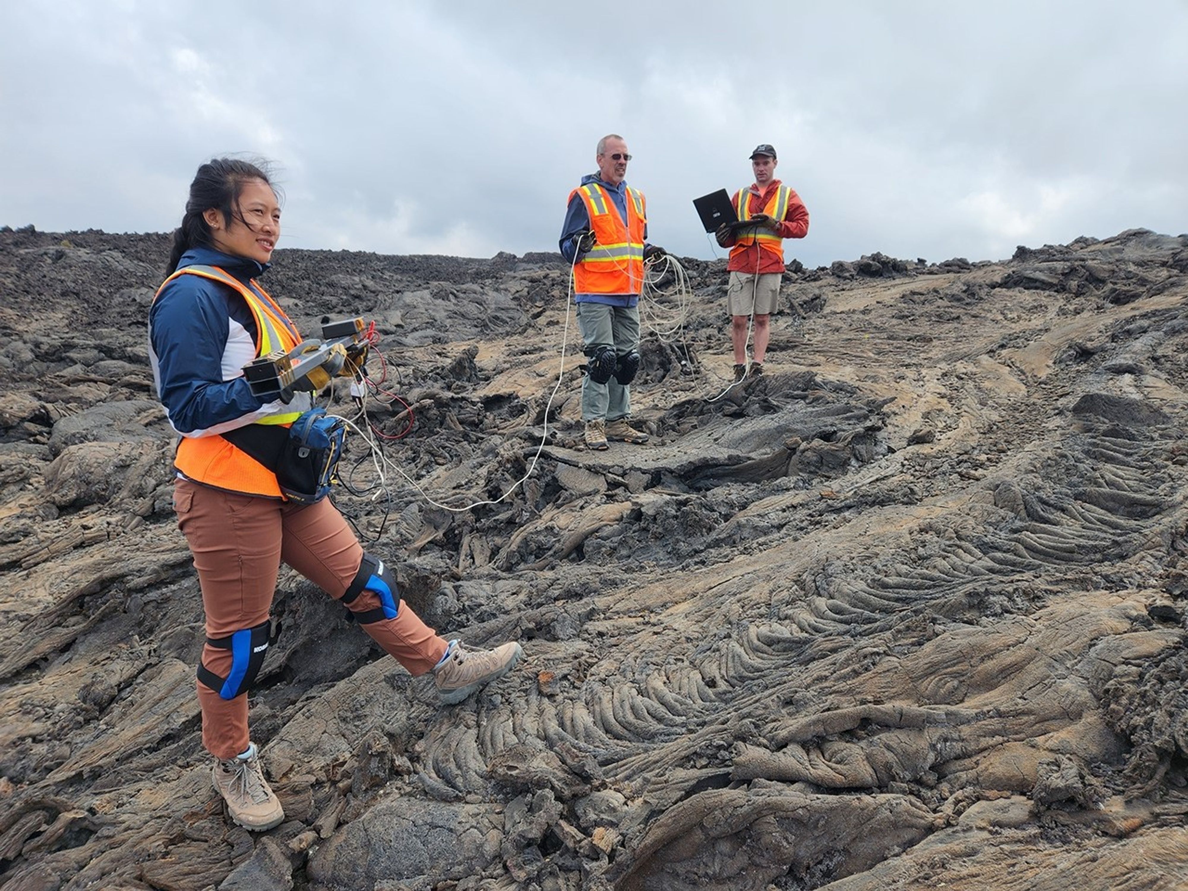 Three people wearing safety vests and standing on a field of lava. The researcher in the foreground holds a metal beam, with electronics attached, in her hands. In the background, two researchers hold power cords and a laptop.