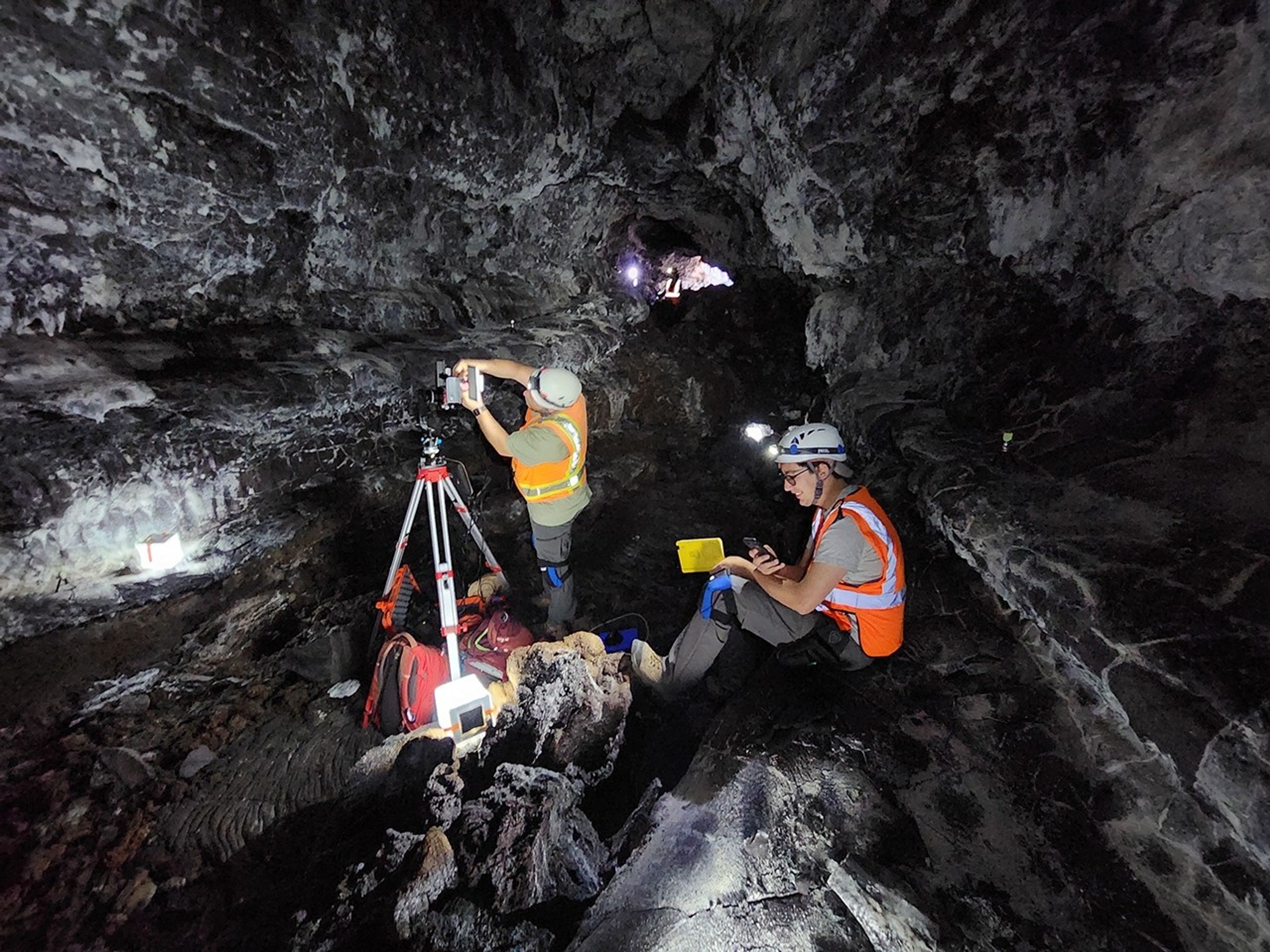 Two researchers wearing high-visibility vests and helmets in a cave. One sits on a natural bench and takes notes. The other sets up a device on top of a tripod.