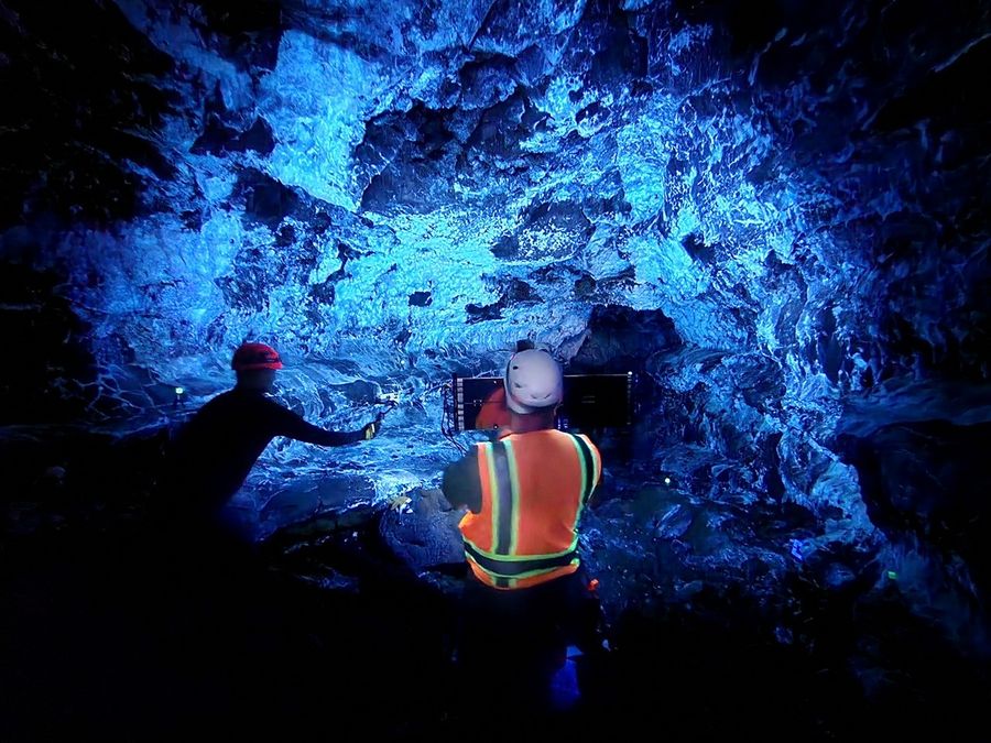 In a blue-lit cave, two researchers wearing helmets operate a large, flat light array mounted on a tripod. The cave walls are coated in a substance that fluoresces brightly.
