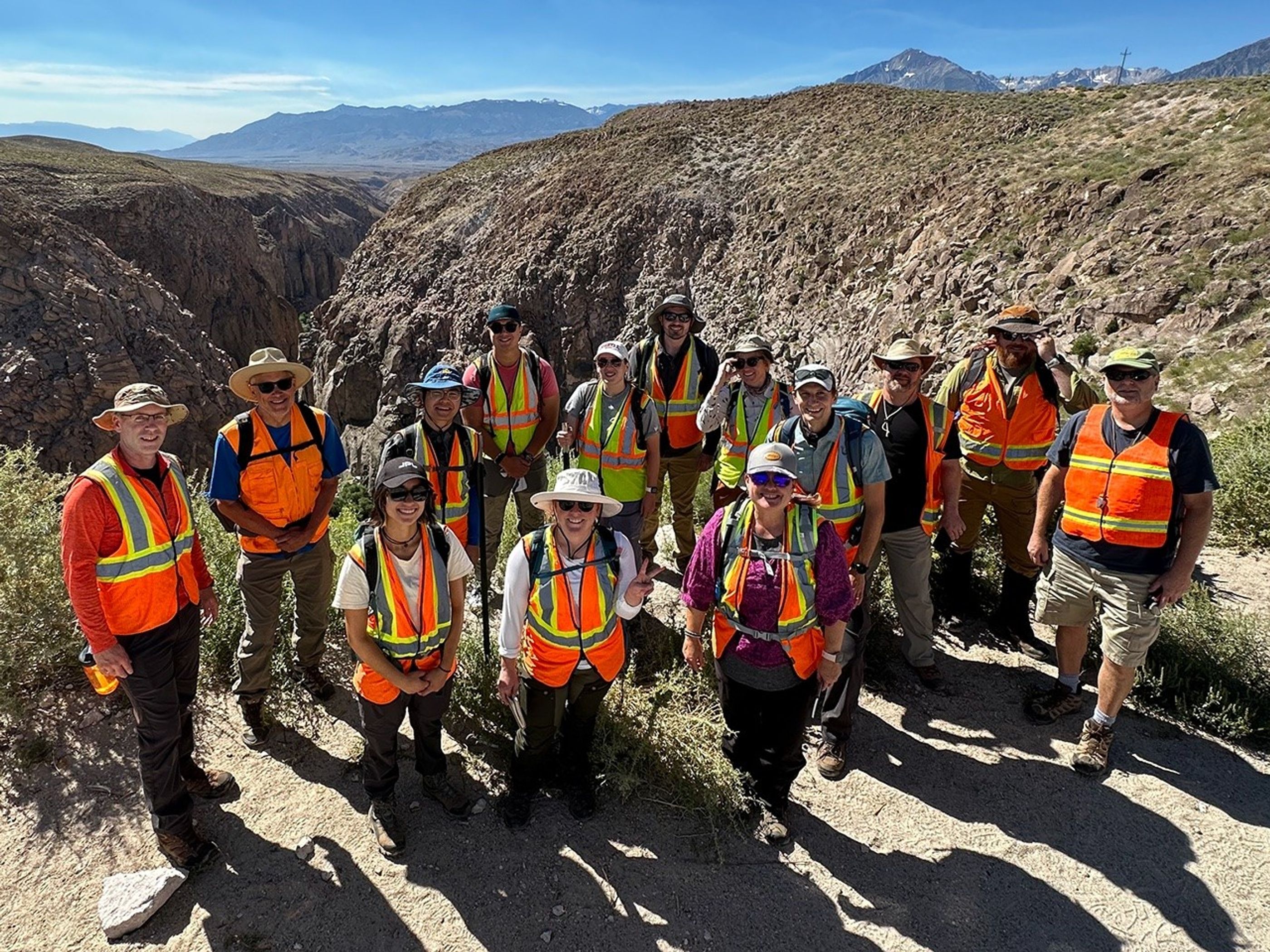 14 smiling people wearing high-visibility vests and sun hats, with a dramatic river gorge in the near background, and mountains on the distant horizon. The researcher in front and center of the group raises her hand with two fingers extended in a peace sign.