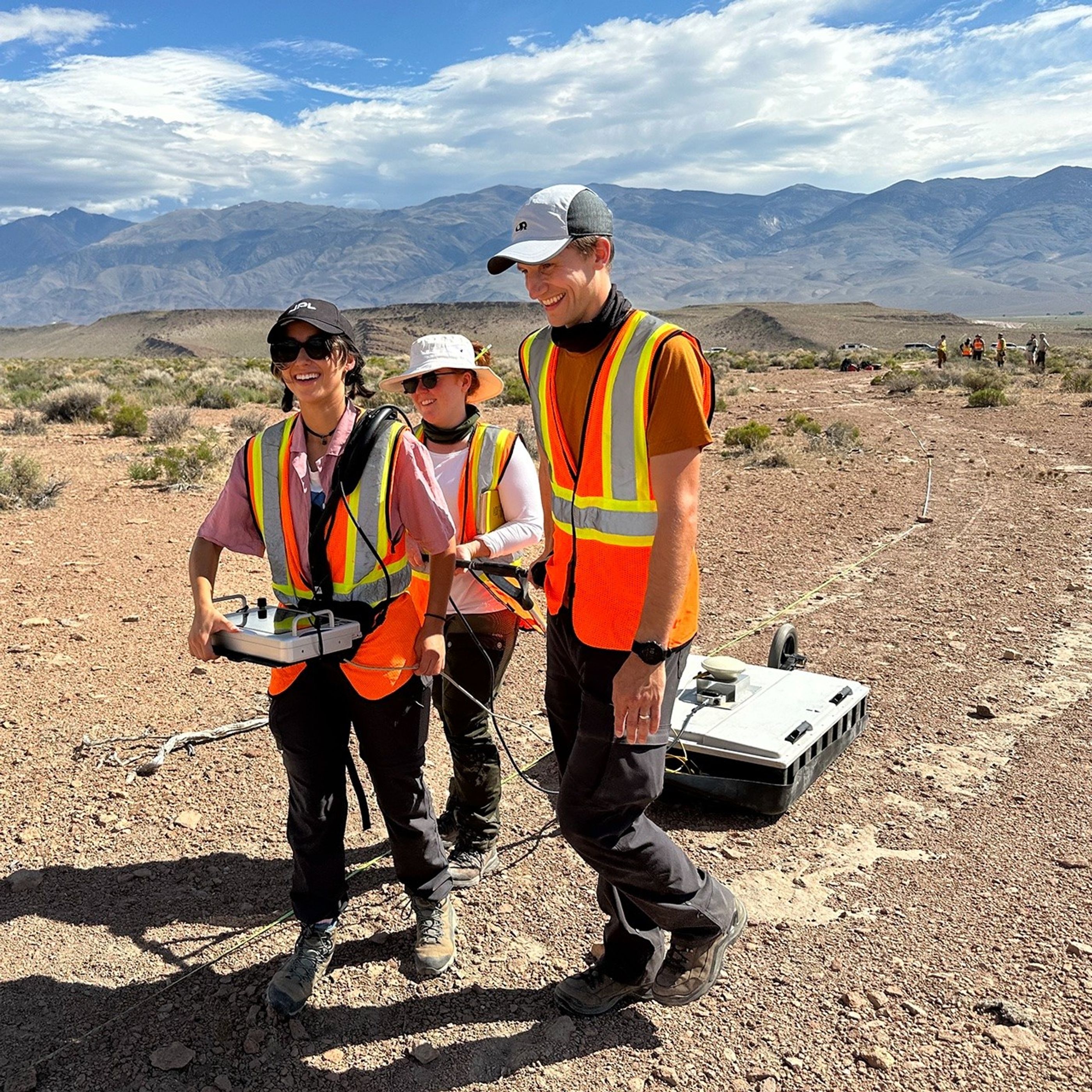 Three smiling people, wearing sun hats and high-visibility vests, at work in the desert. One researcher wears a harness attached to a box with cables coming out, and holds the edges of the box up in her hands. Two use a large handle to pull a flat, rectangular apparatus along behind them. Small shrubs are in the near background, and tall mountains are on the horizon. The sky is blue with white clouds.