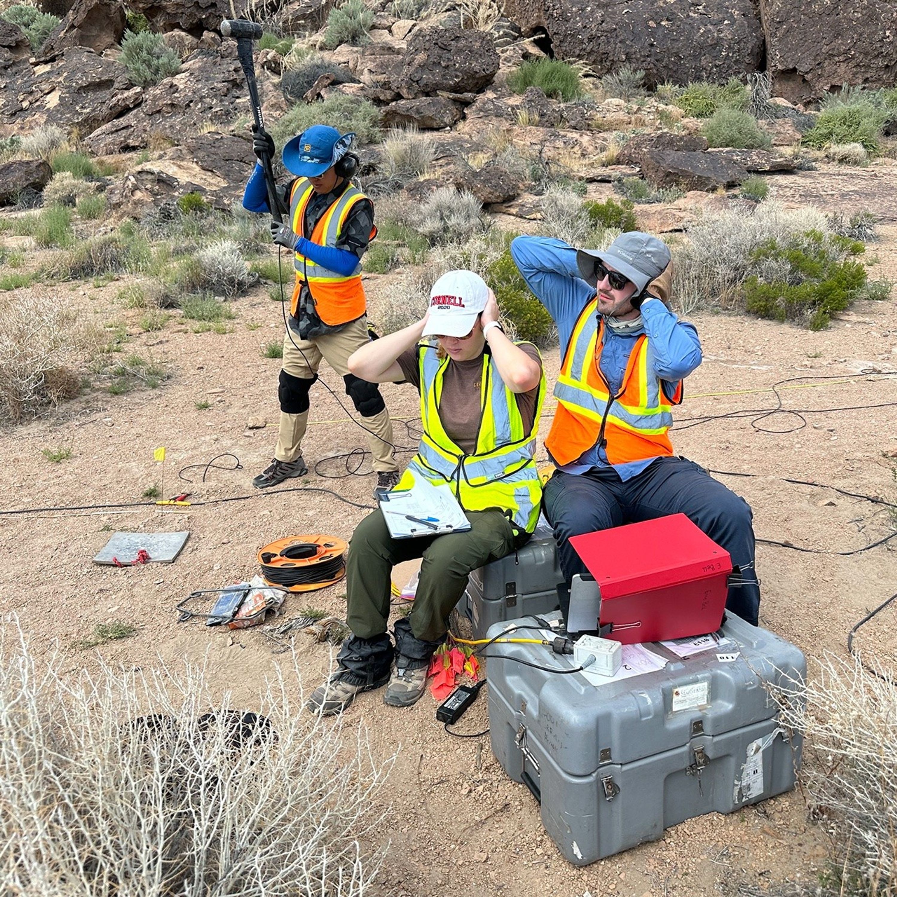 Three researchers wearing bright colors in a rocky, shrubby desert. The person in the background swings a large mallet towards a metal square on the ground. The two people in the foreground, sitting on boxes of equipment and looking at a laptop shaded by a three-ring binder, cover their ears.