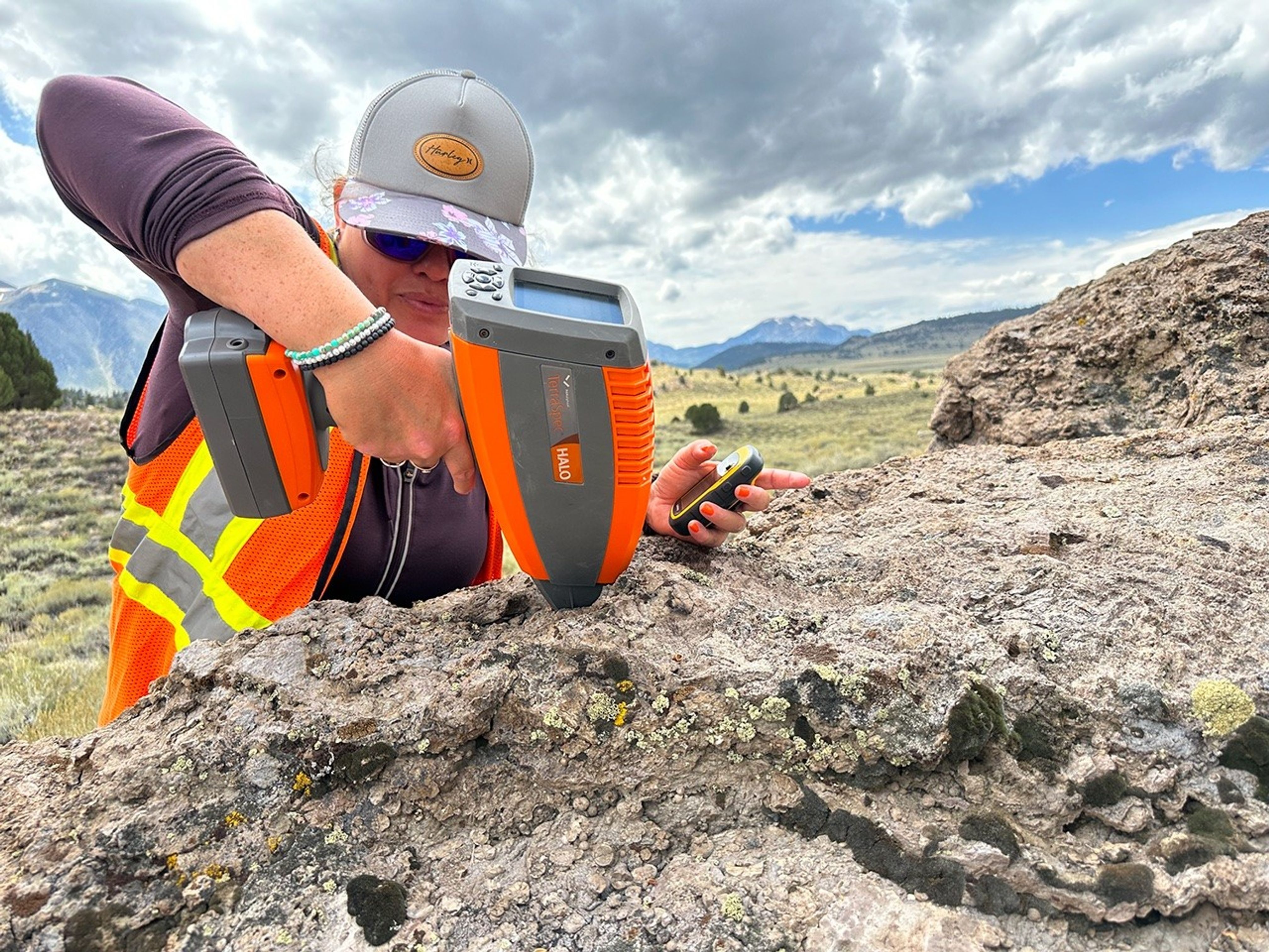 A researcher wearing a high-visibility vest and purple shirt uses one hand to place a portable science instrument against a rock outcrop. The device is orange and grey and resembles a large hair dryer with a digital readout screen. The scientist holds a small GPS unit in her other hand. Sage and yellow vegetation cover the near background. In the distance, mountains rise under a cloudy sky.
