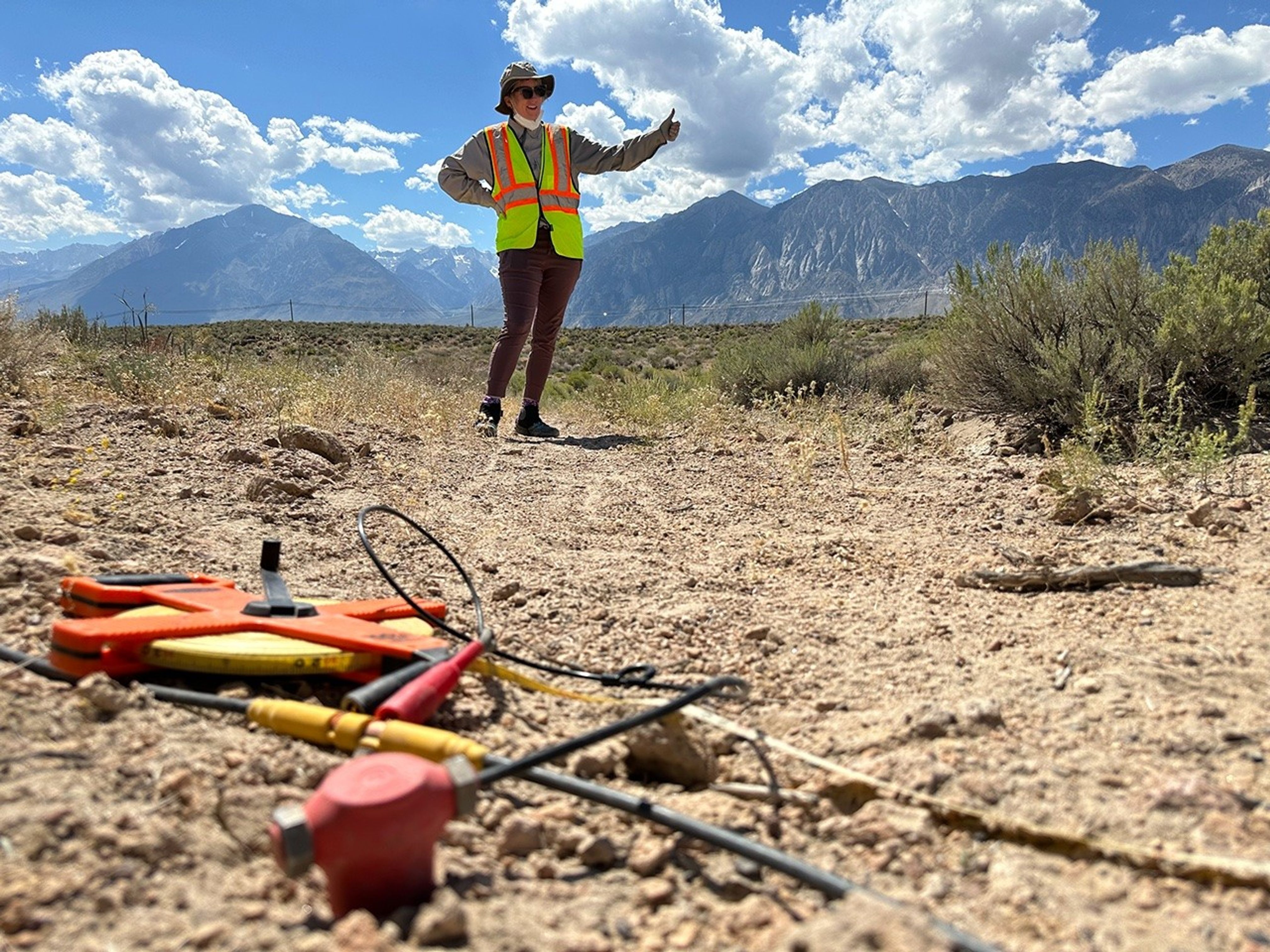 In the foreground, a small red plastic and metal object is driven into the desert ground next to a tape measure. A person wearing a high-visibility vest gives a thumbs-up. Blue mountains line the horizon behind her.