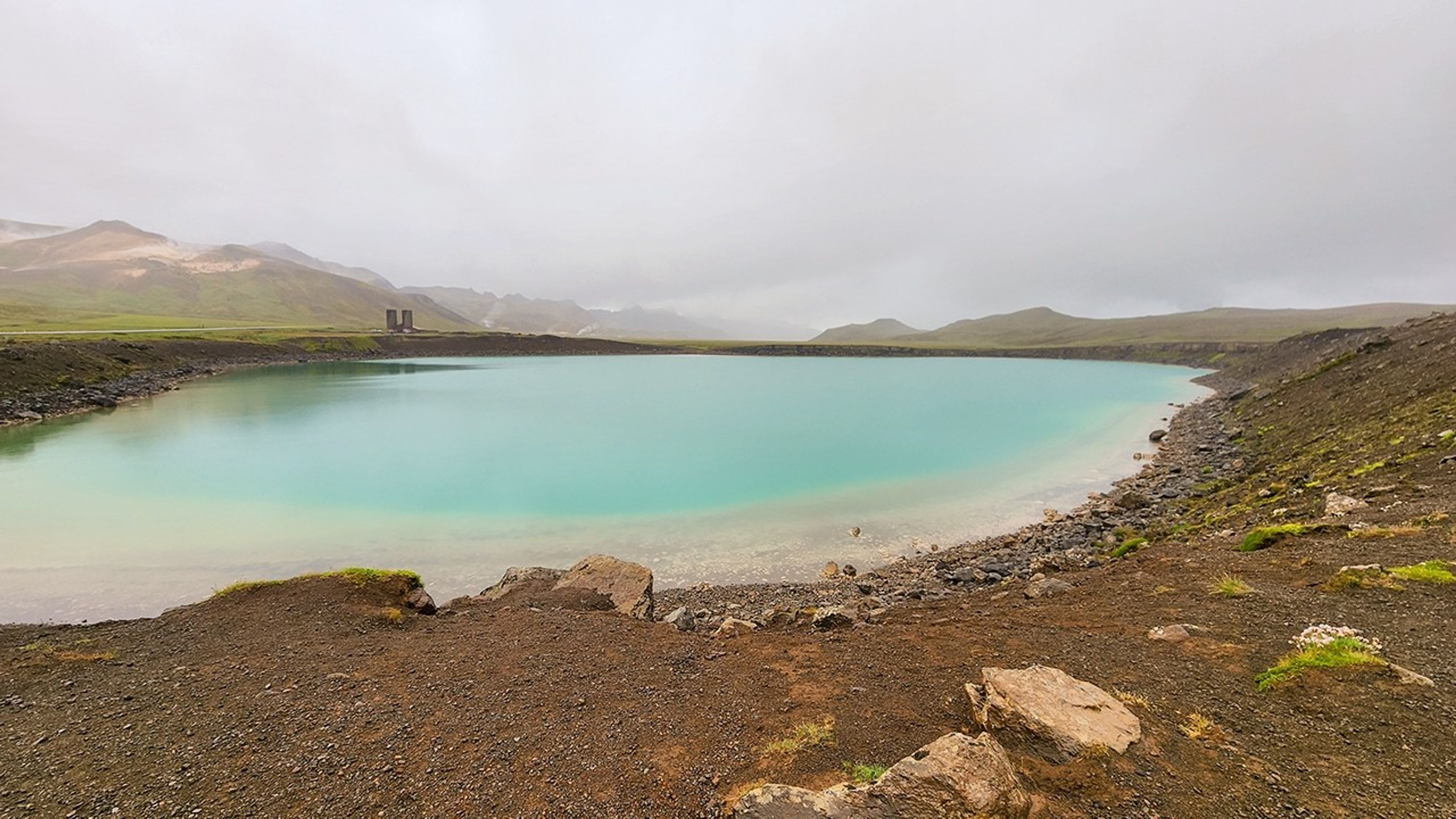 A turquoise-blue volcanic crater lake surrounded by dark volcanic rock shores and moss-covered terrain under overcast skies in Iceland's geothermal landscape.