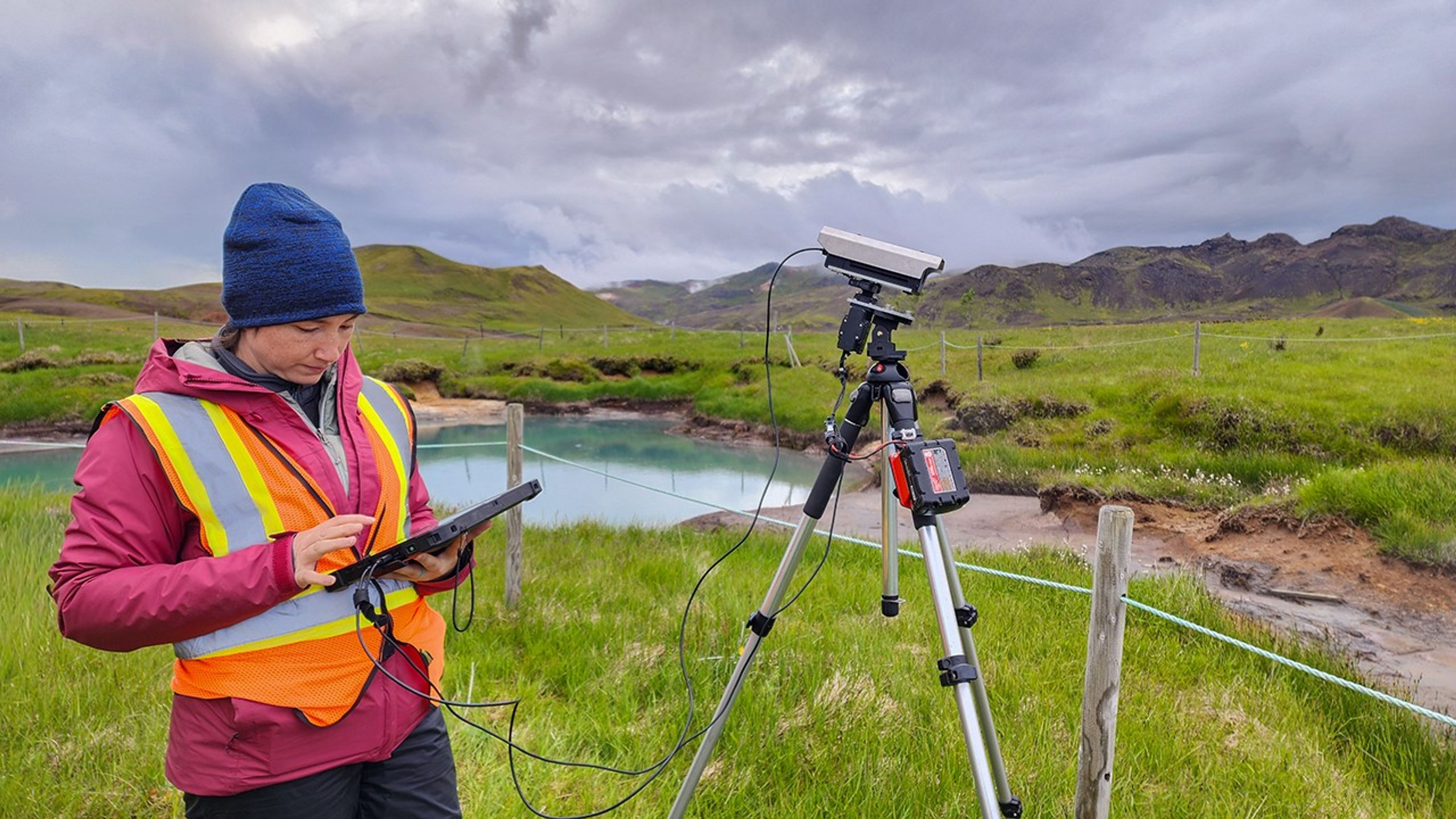 A scientist in a safety vest operates a hyperspectral imaging instrument on a tripod, with a tablet controller connected by cable, overlooking a turquoise geothermal pool.