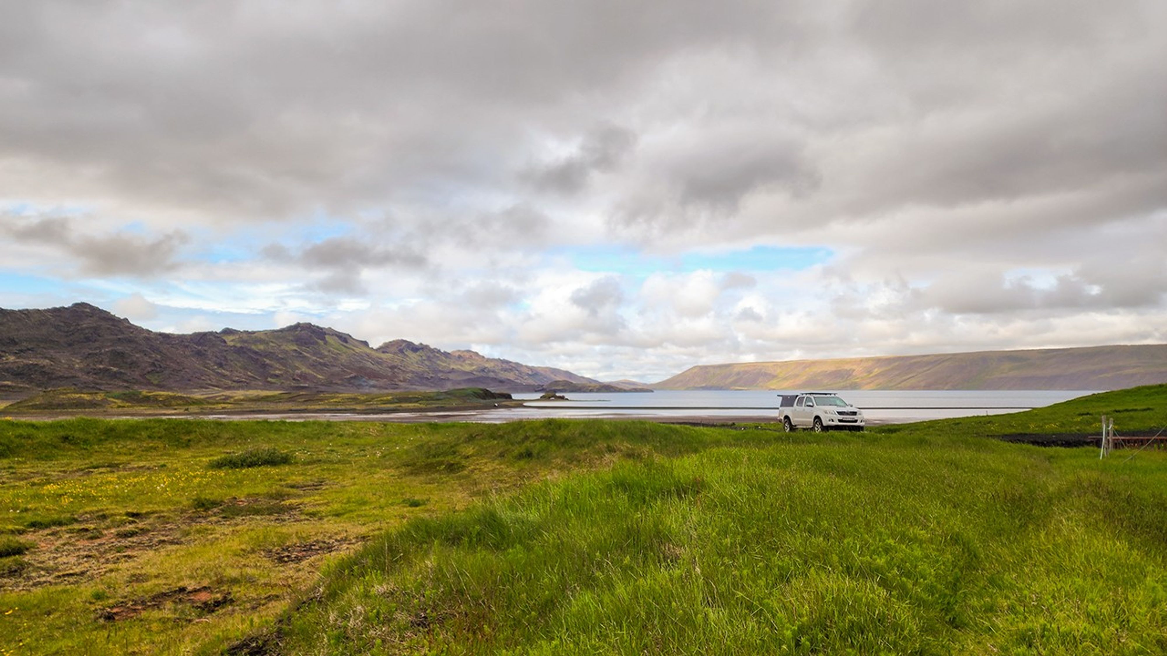 A white SUV with roof rack parked beside Lake Kleifarvatn, with vibrant green grass in the foreground and dramatic volcanic mountains reflected in the calm water under cloudy skies.