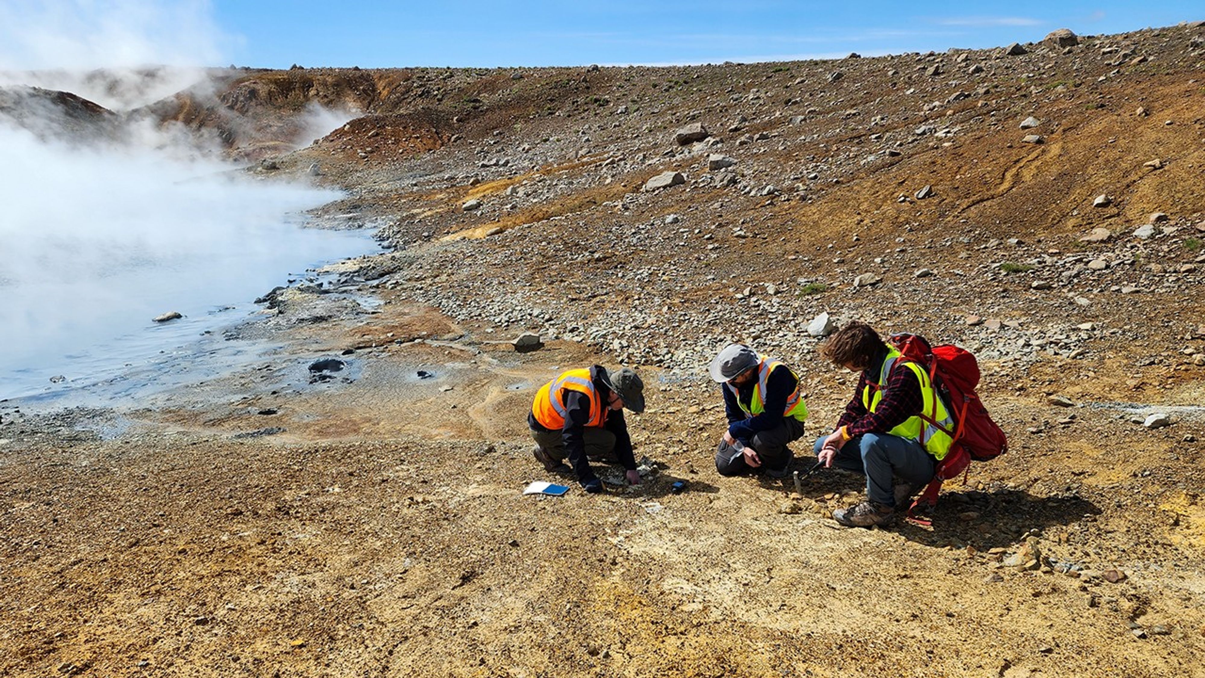 Three scientists in safety vests crouch on yellow-orange sulfur-stained ground near a steaming geothermal vent, collecting samples on the slope of a volcanic hillside.