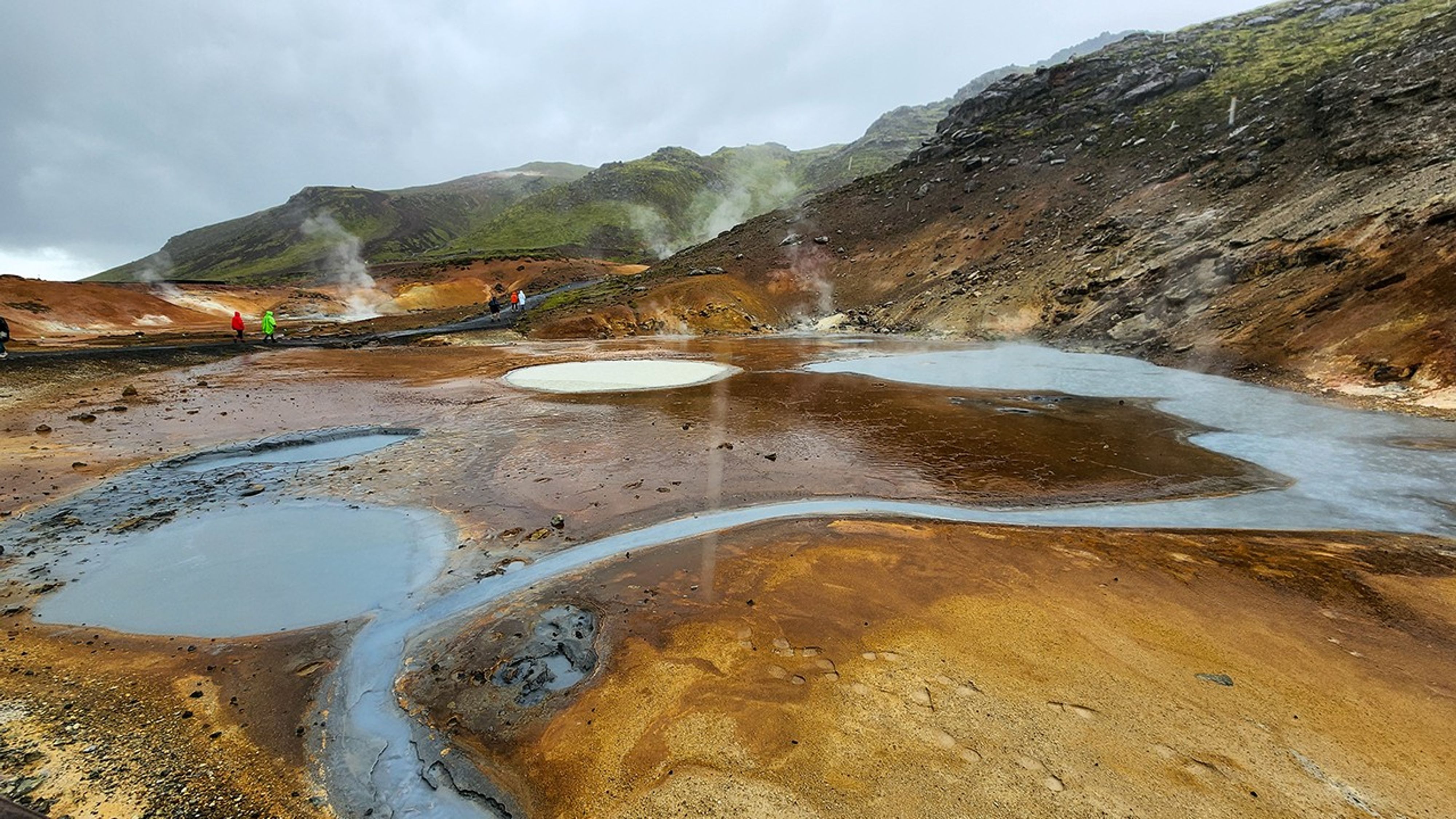 Colorful geothermal landscape showing multiple steaming pools with blue, gray, and orange mineral deposits, mudpots with concentric patterns, and research team members visible in the distance.