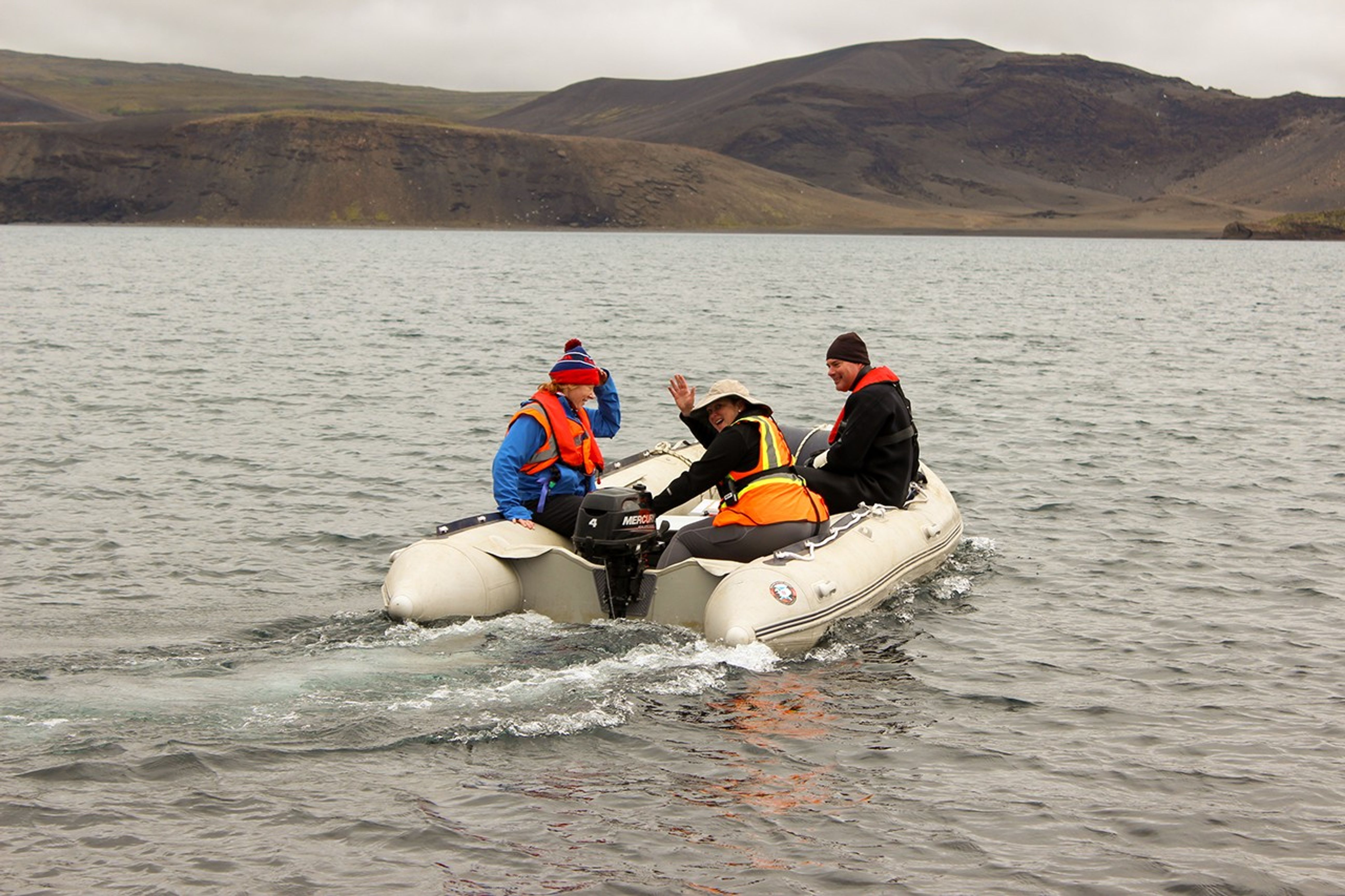 Three team members in life jackets aboard an inflatable motorboat on Lake Kleifarvatn, with one waving to the camera and barren volcanic hills visible across the water.
