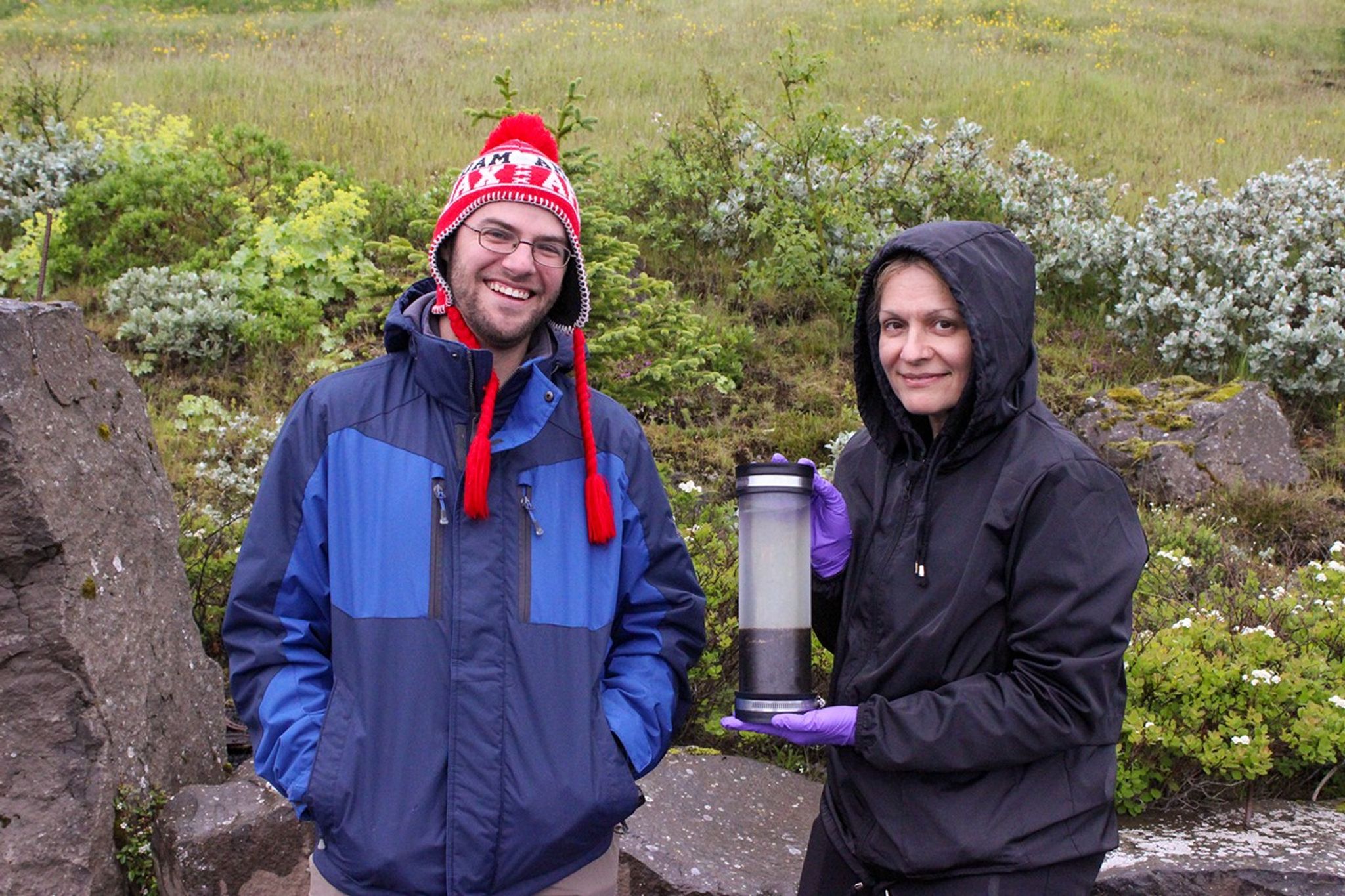 Two scientists in winter gear hold up a cylindrical gas sampling device, standing among moss-covered lava rocks and white flowering plants in Iceland's volcanic terrain.