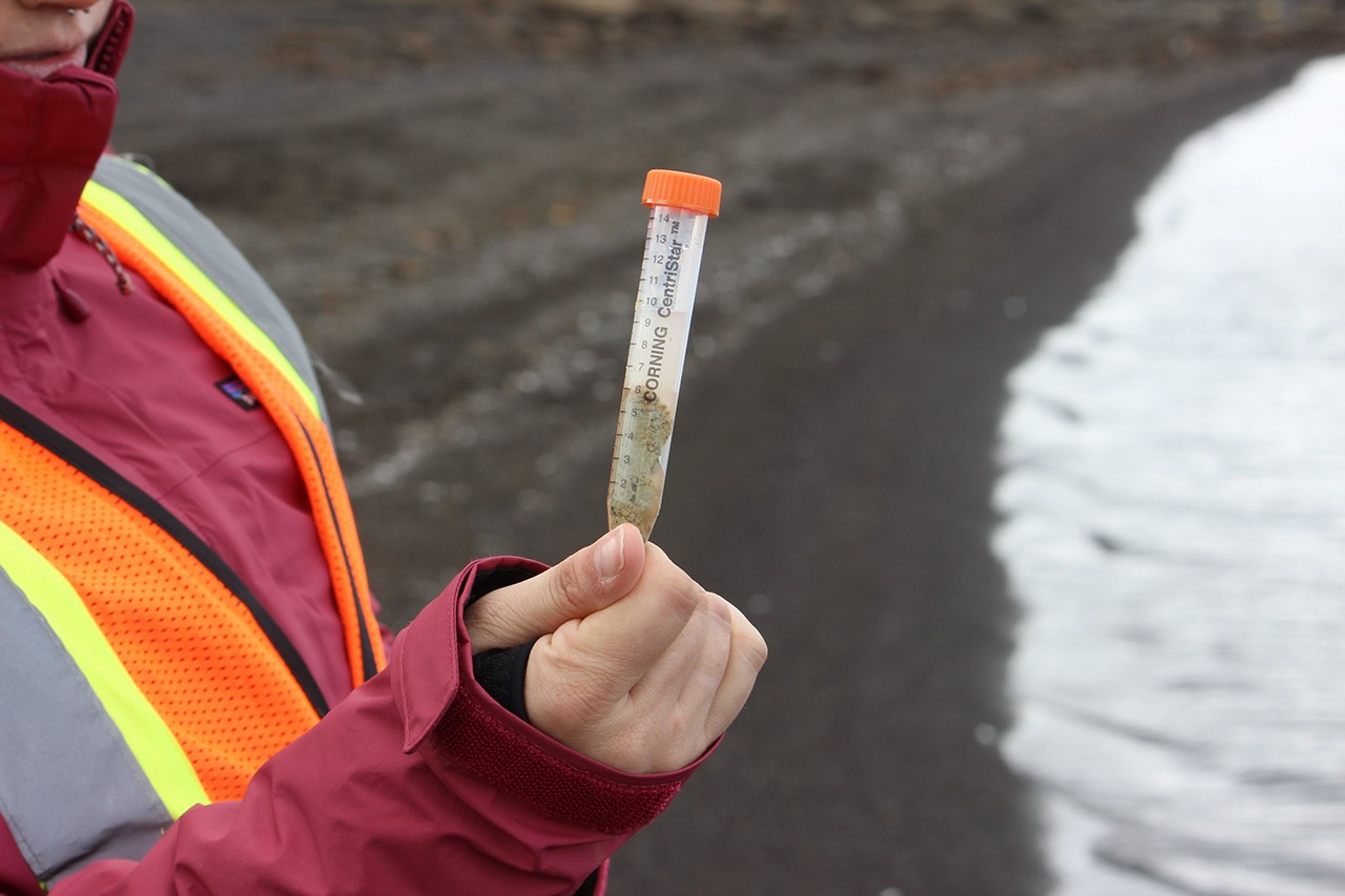 A researcher in a safety vest holds up a labeled centrifuge tube containing sediment and water sample collected from a geothermal pool or lake edge.