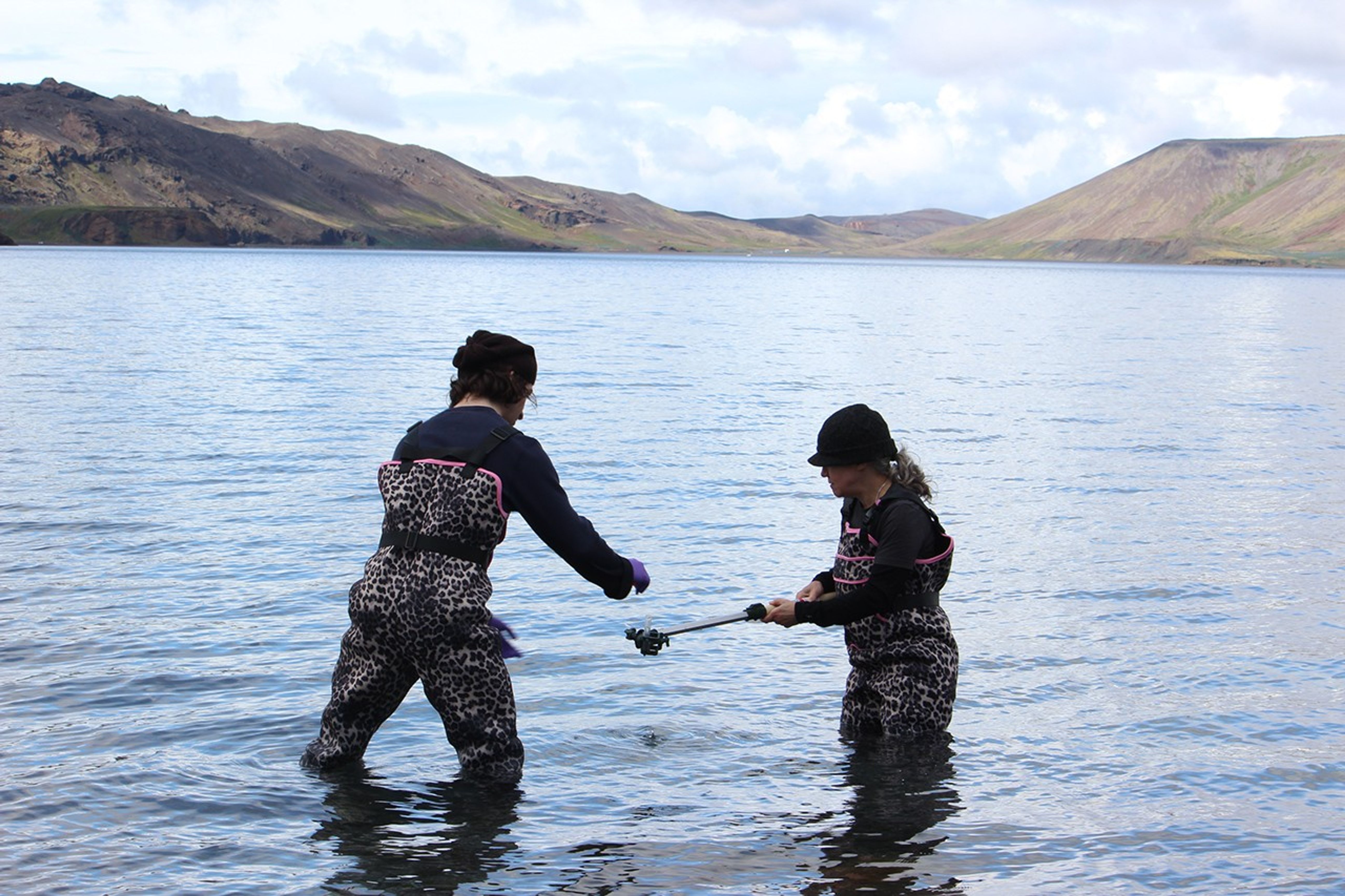 Two scientists in cheetah-print waders work together in shallow lake water, using a long pole to collect samples from the lake bottom under sunny skies.