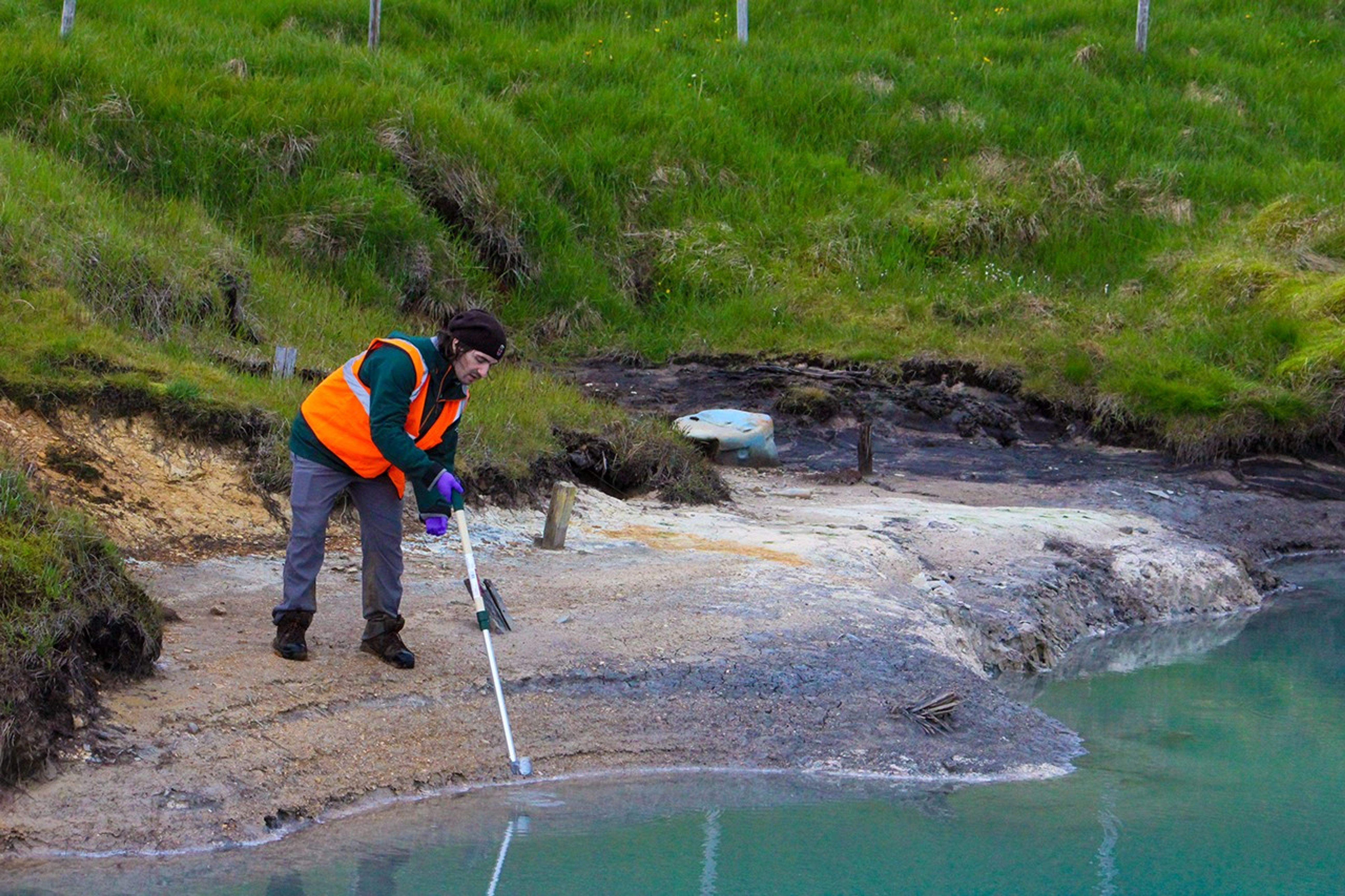 A person in an orange vest uses a long rod to take a sample from the lake. The lake is a turqoise green color. In the background there is bright green grass.