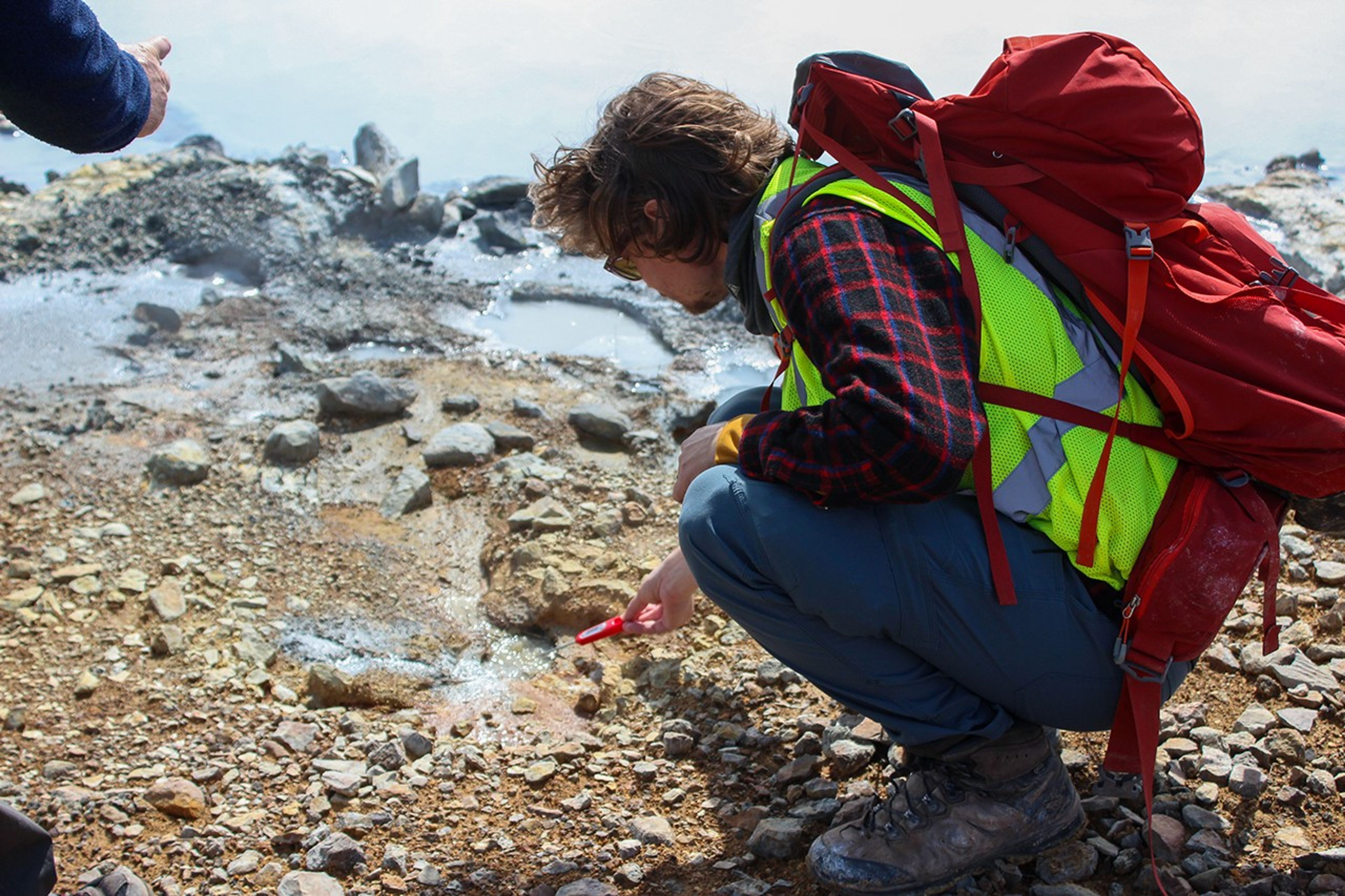 A man wearing a green safety vest, red plaid shirt, blue pants, brown boots, and a red backpack crouches down to take a temperature of boiling water near a geothermal vent. In the background, the fluid is grey and pools in small amounts. In the foreground, the dirt is an orange brown and speckled with rocks.