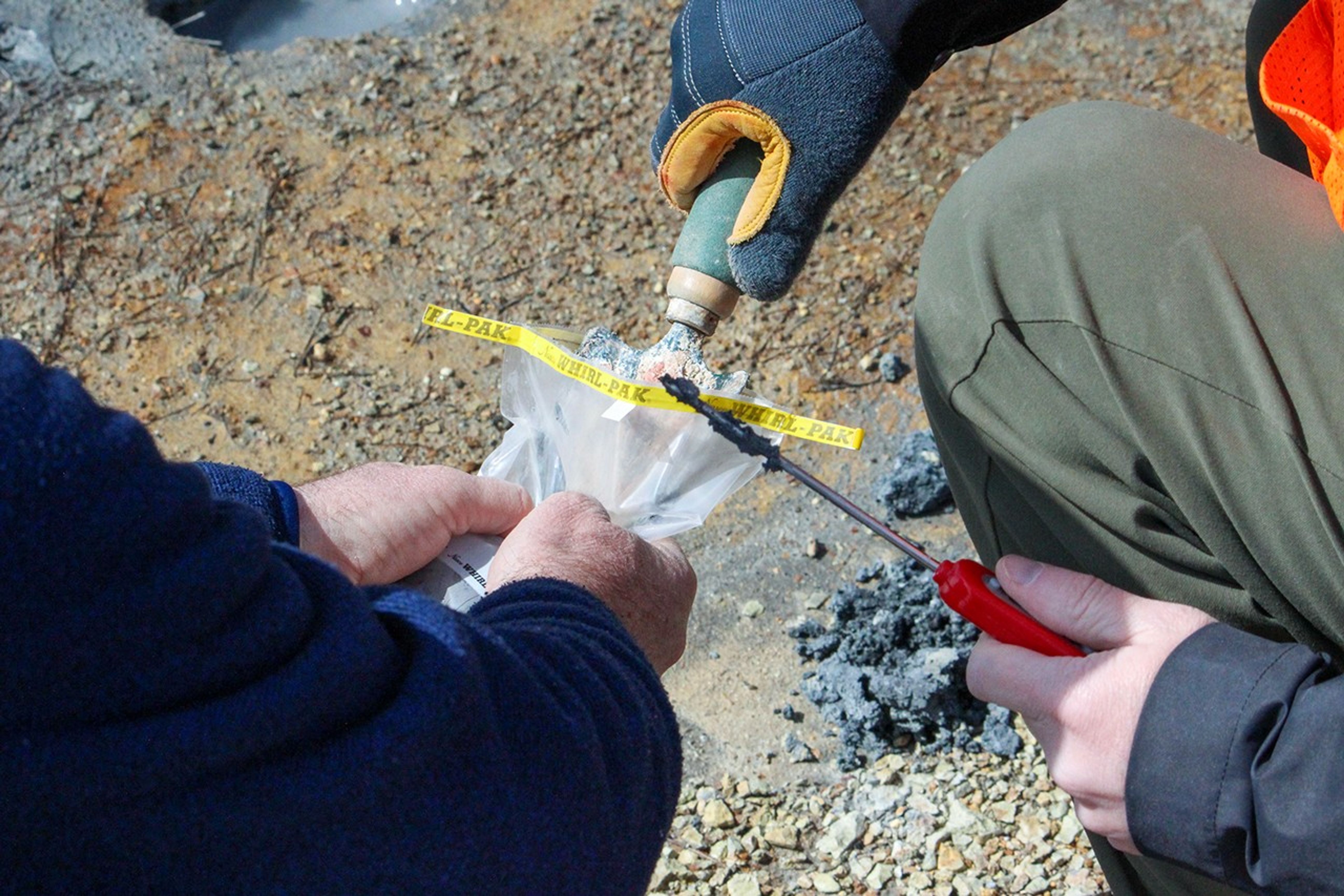 Scientists collect a geothermal sample using a small metal scoop and sample bag marked with yellow field tape on mineral-stained volcanic ground.