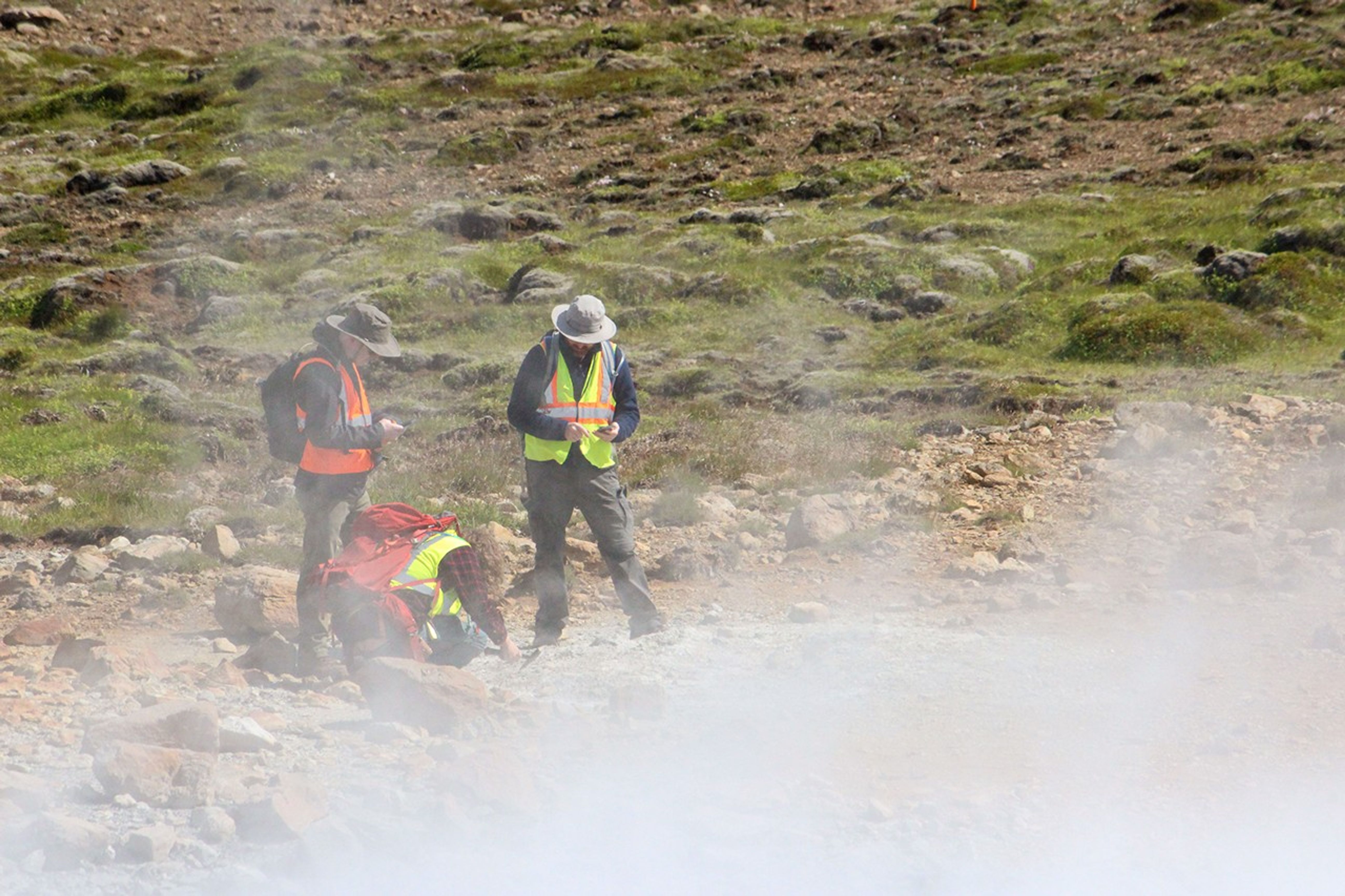 Three researchers work through dense steam from an active fumarole, with their red backpack visible on the ground amid moss-covered volcanic landscape.