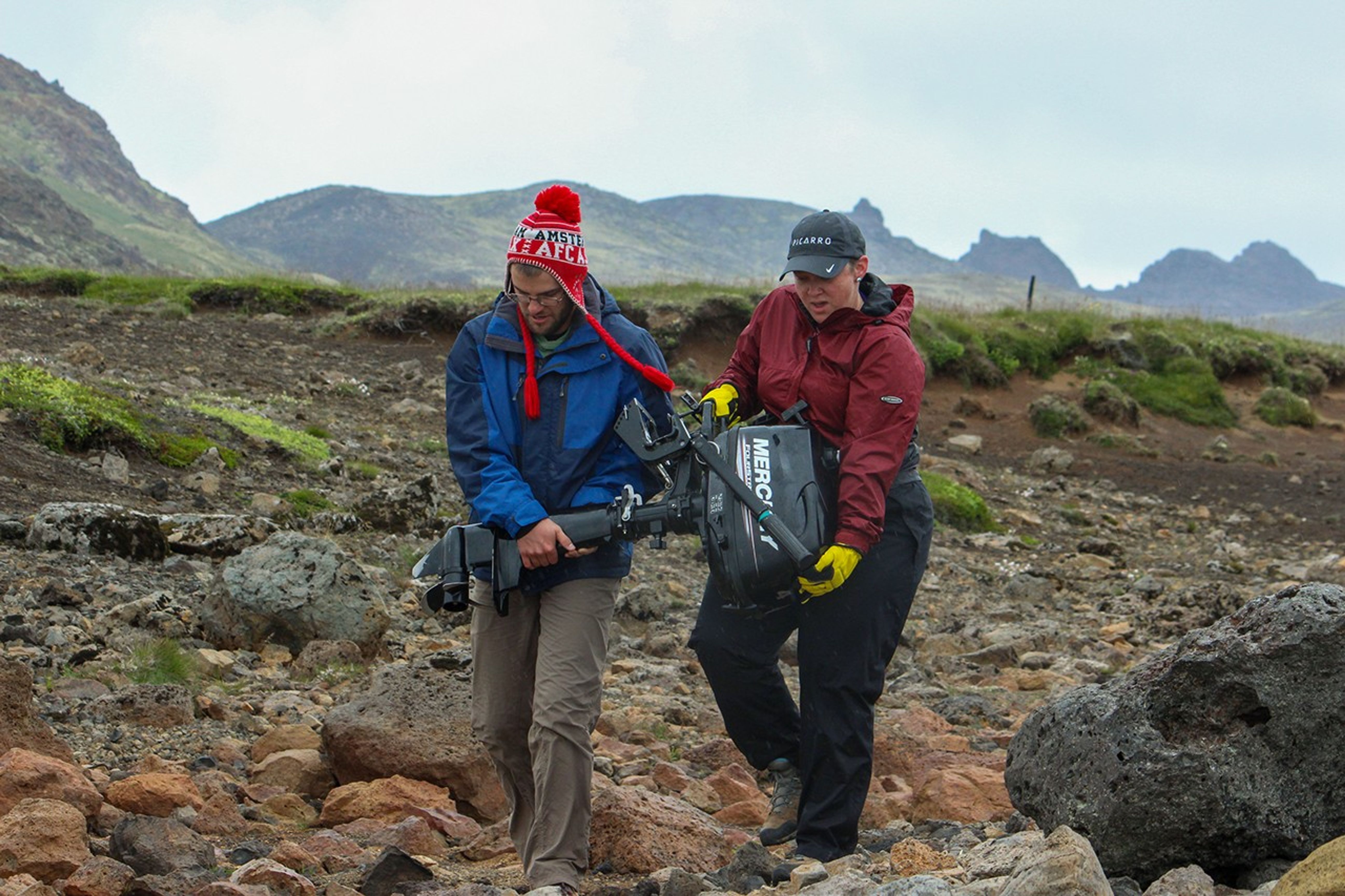 Two scientists carry heavy equipment cases across rocky volcanic terrain with jagged lava formations, walking toward distant mountains under overcast skies.