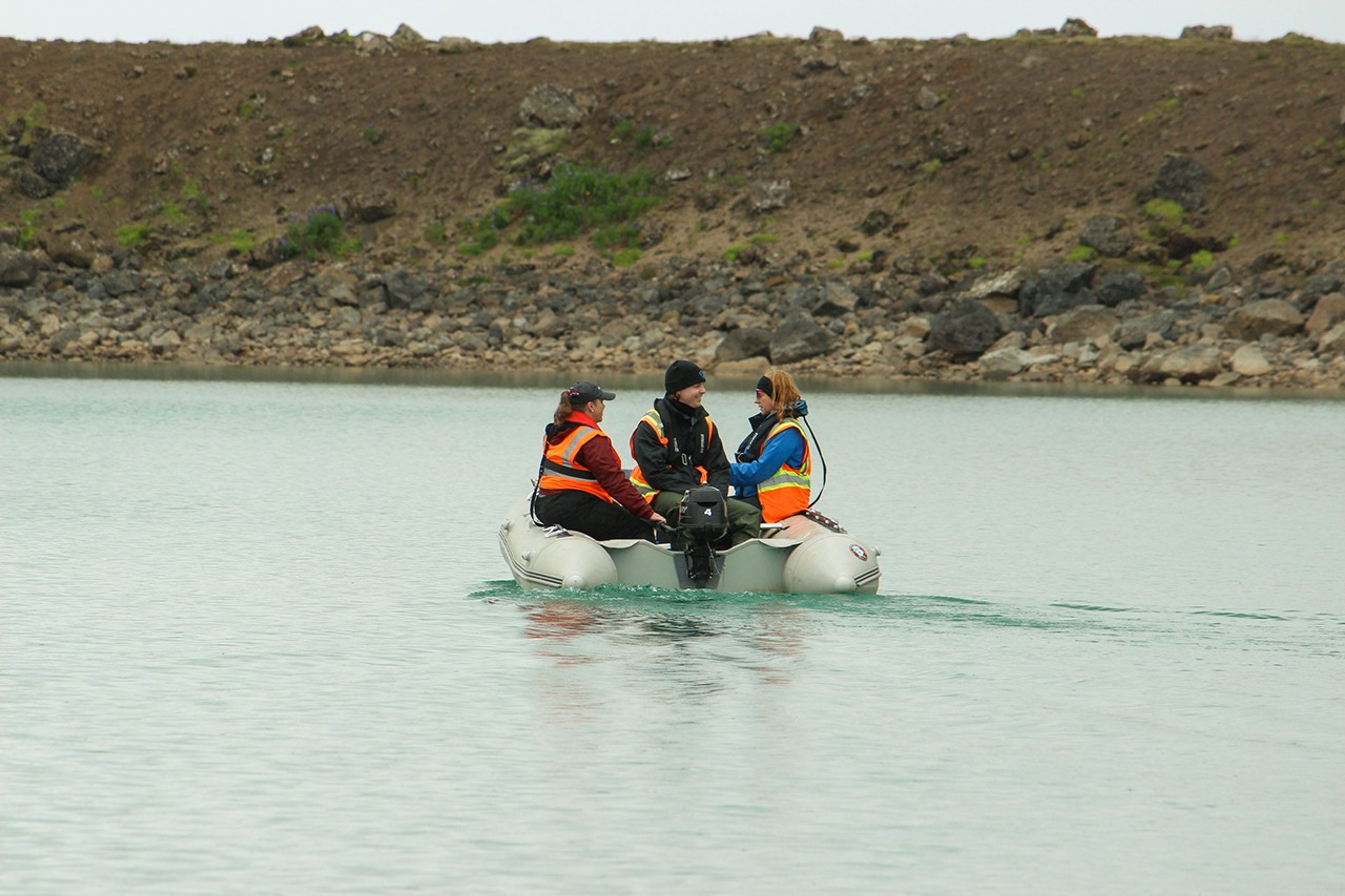Three scientists in life jackets and safety vests sit in an inflatable boat on a turquoise volcanic lake, conducting water sampling with bare volcanic slopes in the background.