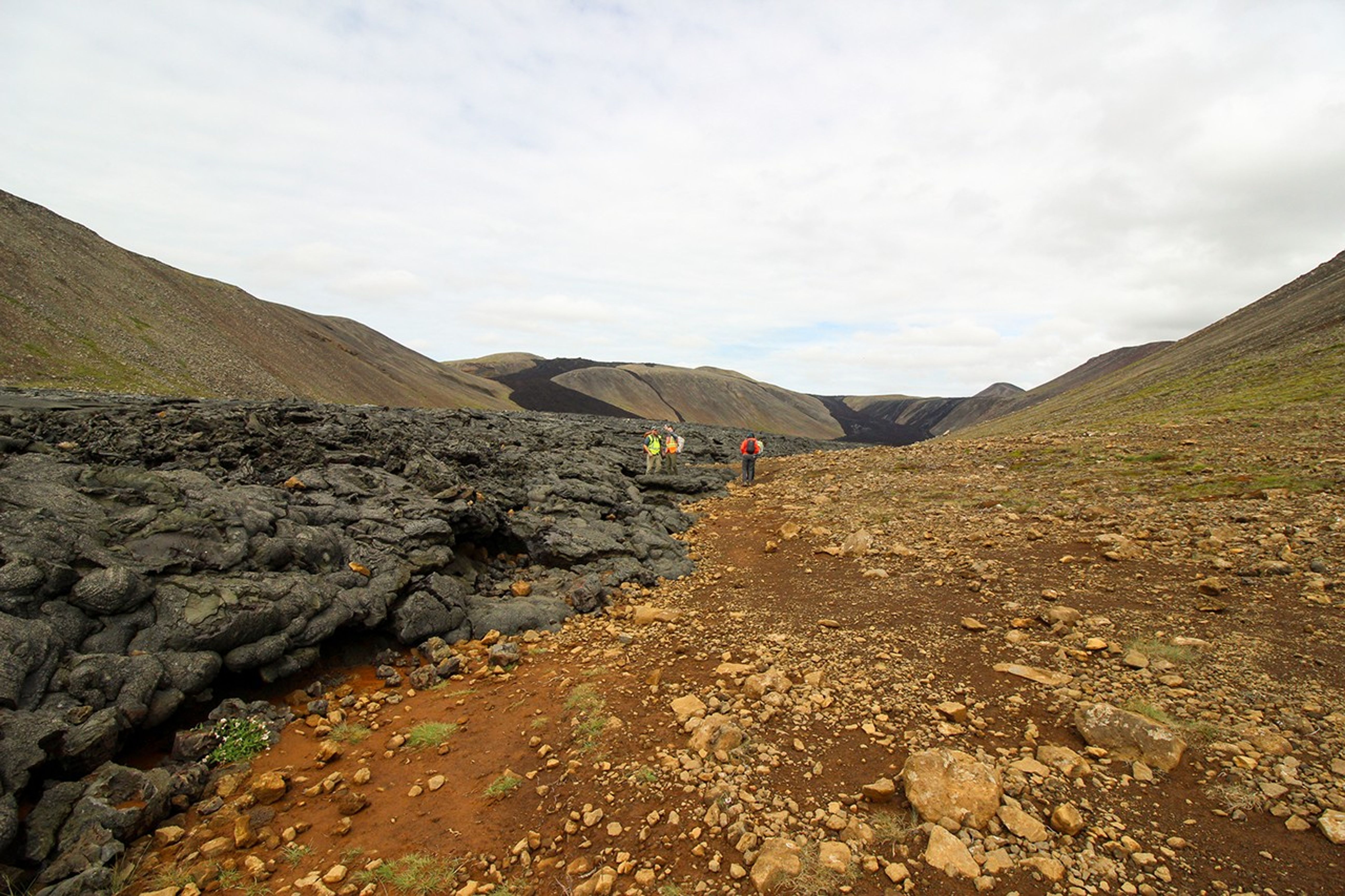 Scientists in bright safety vests hike along the boundary between rough black lava flows and orange-brown oxidized terrain in a volcanic valley.