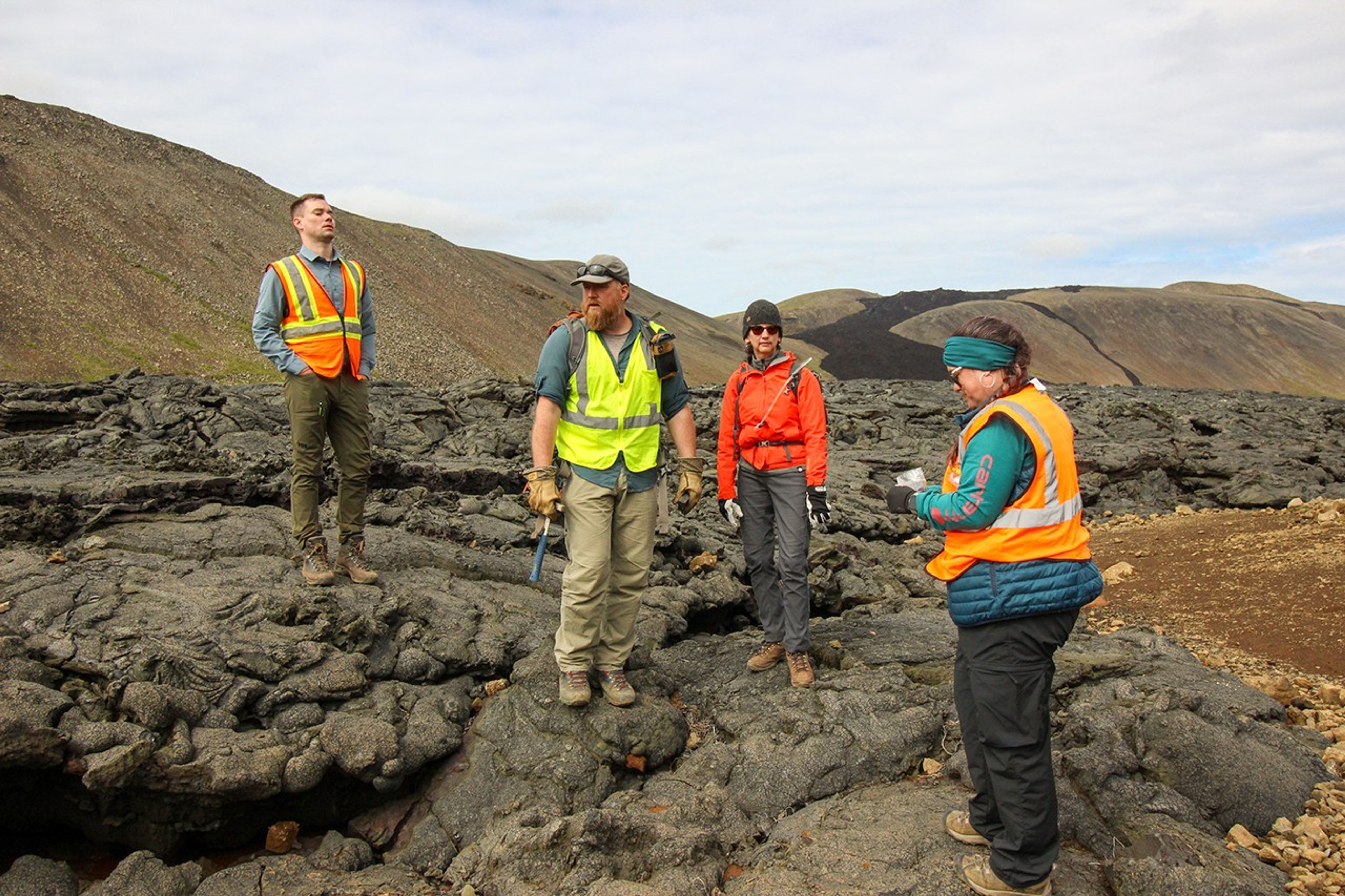 Four team members in safety vests stand on textured black lava rock formations, discussing field observations with volcanic hills visible in the distance.