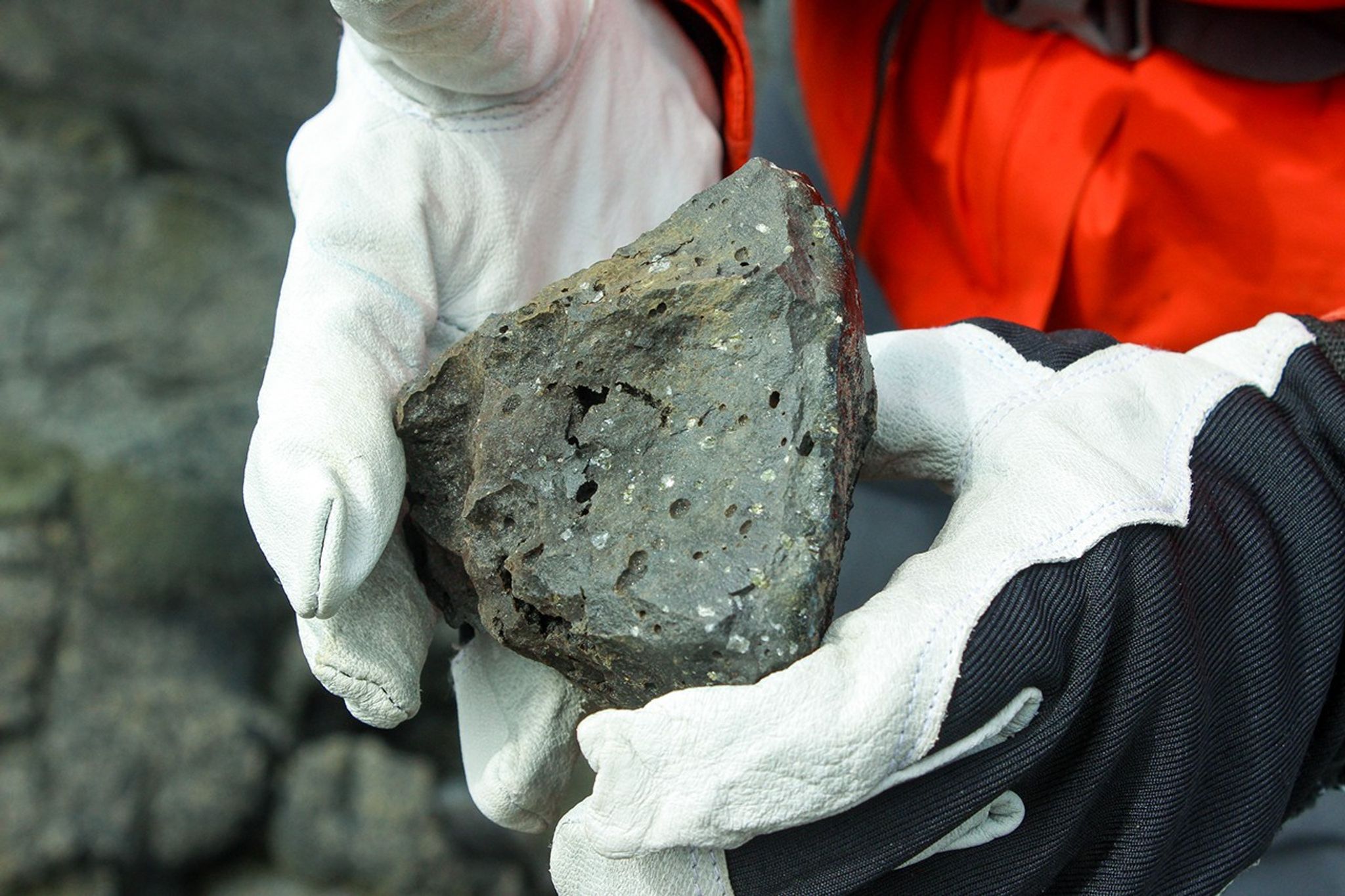 Close-up of gloved hands holding a piece of vesicular basalt showing the porous, bubble-filled texture characteristic of volcanic rock formed from lava.