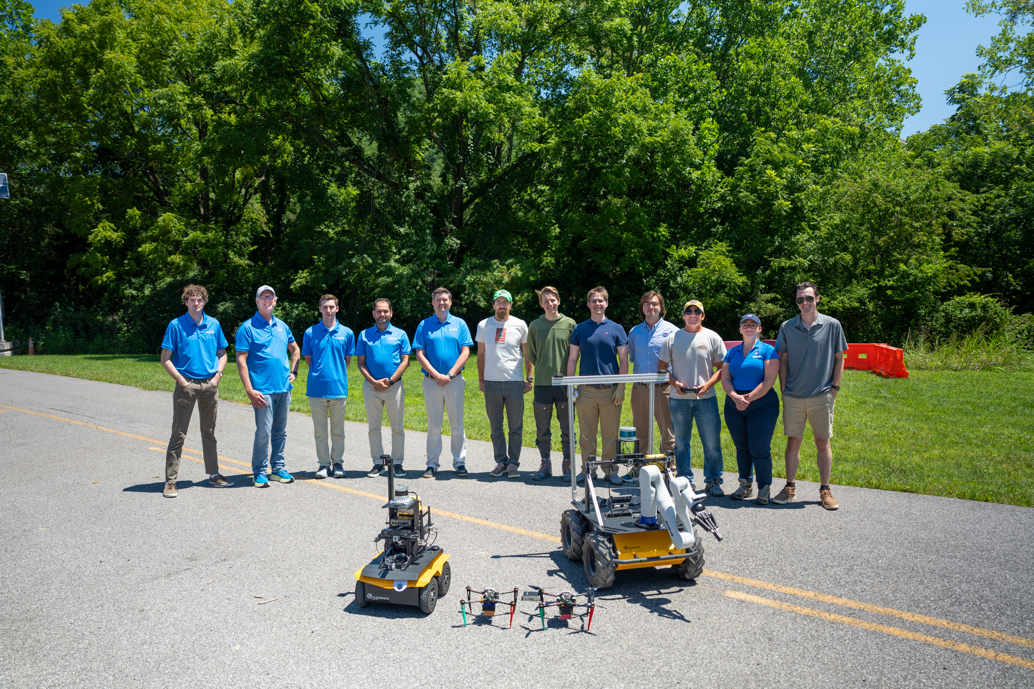 Team photo of twelve people standing on a road with trees behind them. In the foreground are four robots: two rovers and two drones.