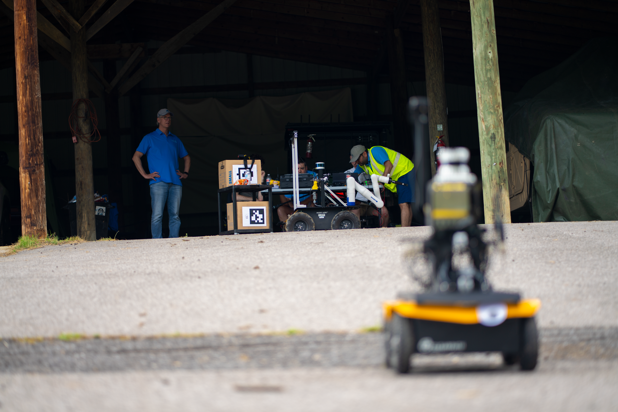 A small robot on the move across pavement. In the background, people observe its progress and work on another robot.