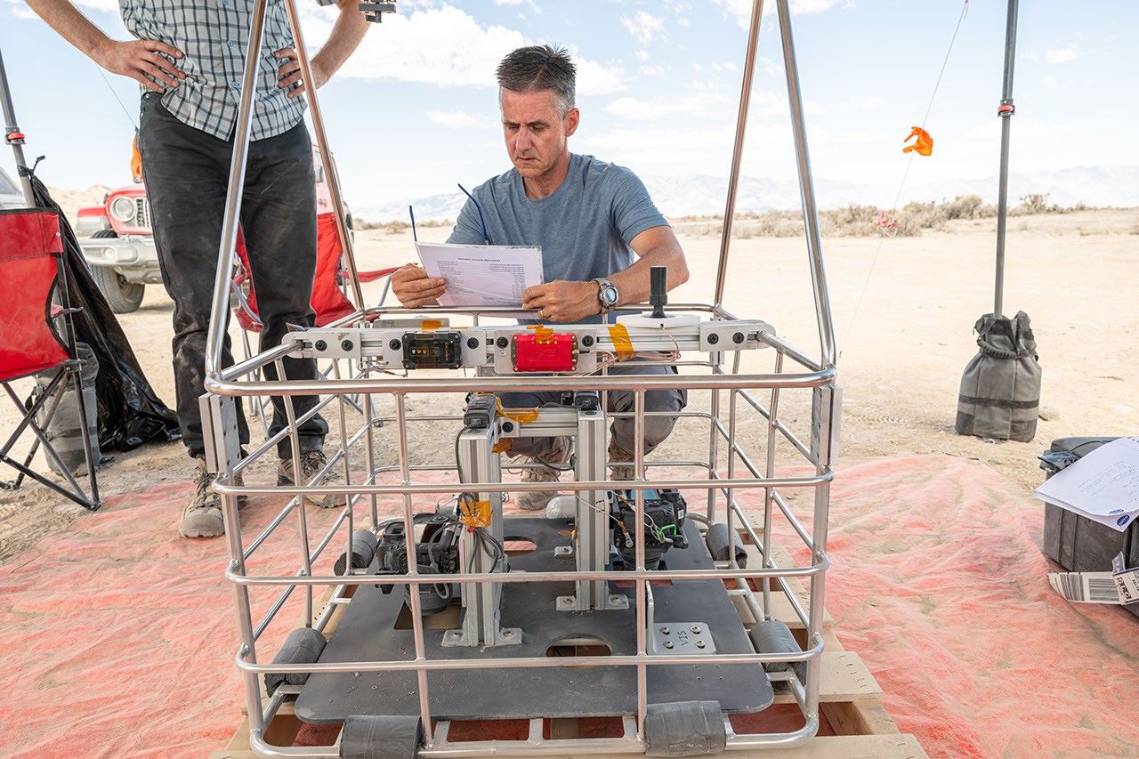 Researcher reviews notes while kneeling beside a square, metal basket of scientific instruments at a desert field site, with another team member standing nearby.