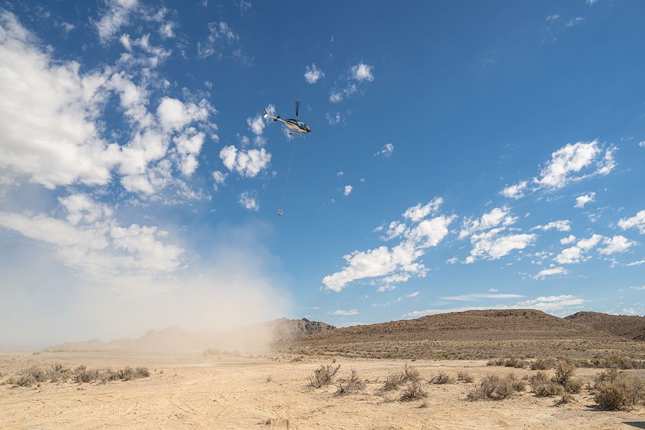 Helicopter lowers equipment by long cable over a dusty desert landscape, creating a large cloud of dust.