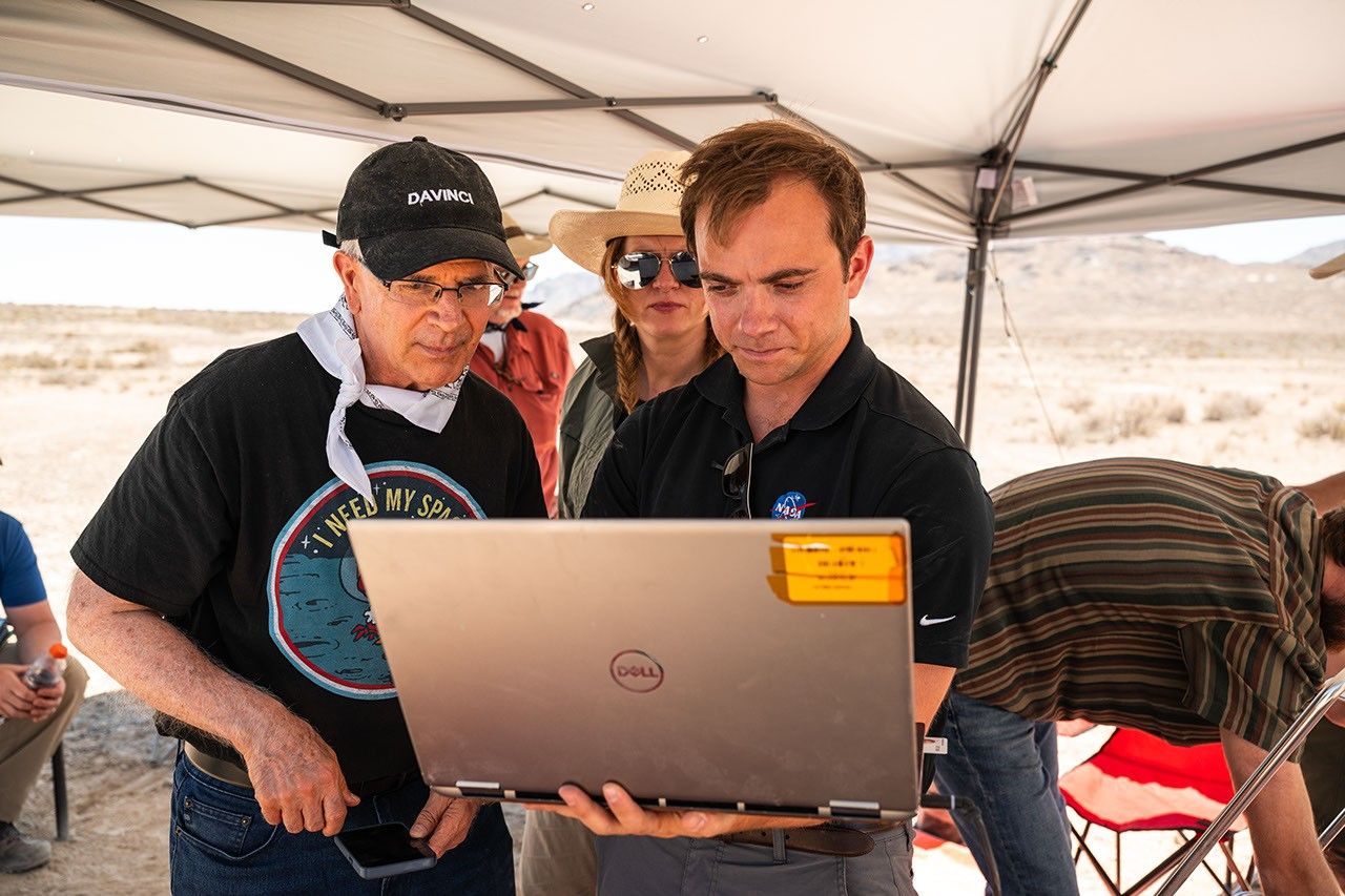 Researchers gather under a shade canopy in the desert, reviewing data together on a laptop during a field science operation.
