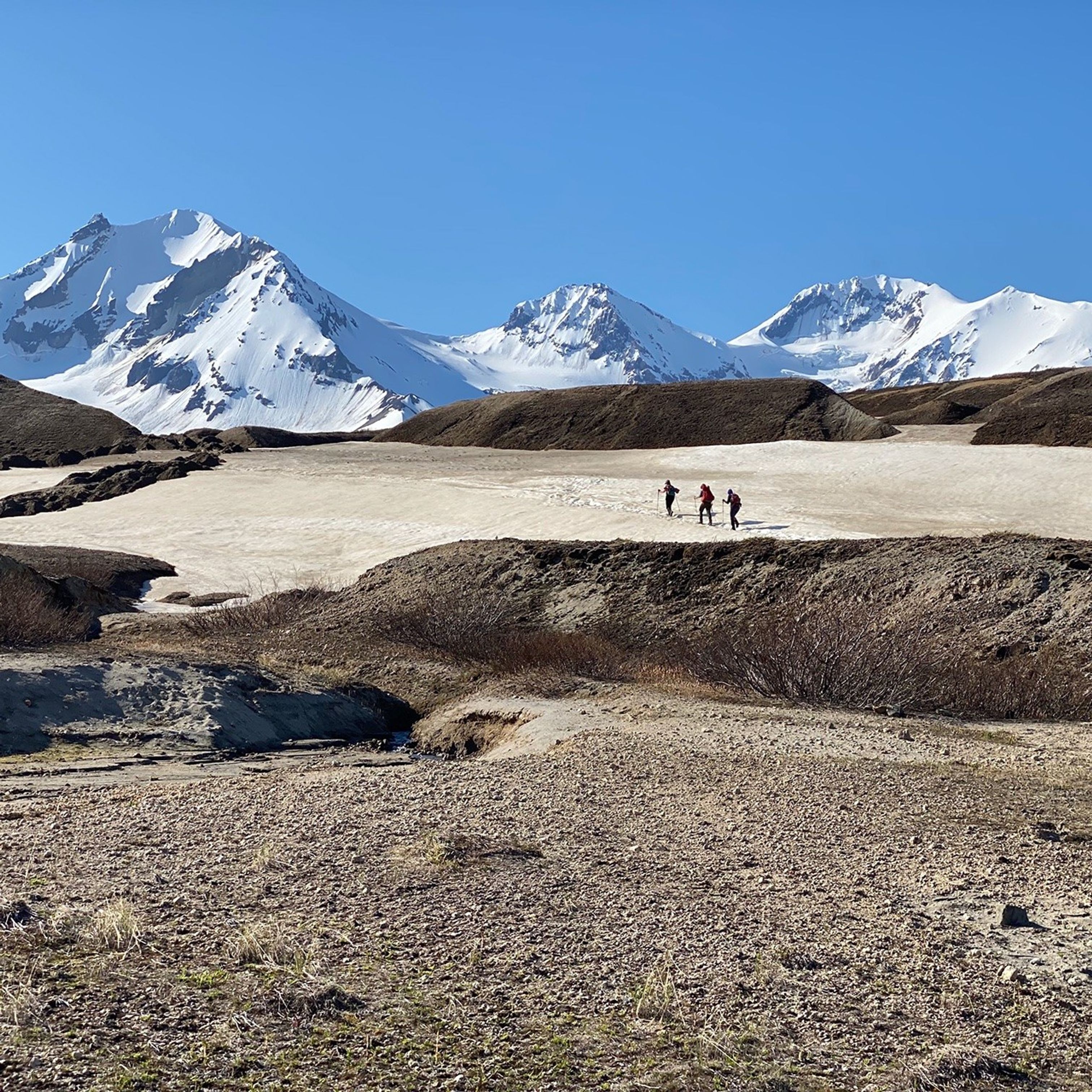 Three people, wearing large backpacks, trek across a snow field between hills of dark rubble. In the background: steep, snow-covered mountains under a blue sky.