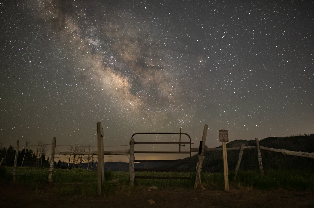 The bright, dusty band of the Milky Way galaxy stretches across a star-filled night sky. In the foreground, a rustic wooden fence and a closed metal gate stand on a grassy field, with dark hills silhouetted in the distance.
