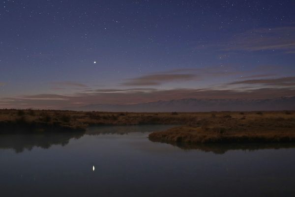 A night landscape shows a bright, reflective river snaking through a flat, grassy plain under a sky full of stars. In the center, Jupiter shines prominently and is reflected in the still water below. The horizon features distant, shadowed mountains, and the sky above them shows faint hints of a reddish glow from the setting or rising sun, possibly indicating twilight. The overall scene is serene and emphasizes the vastness of the night sky and the natural world.