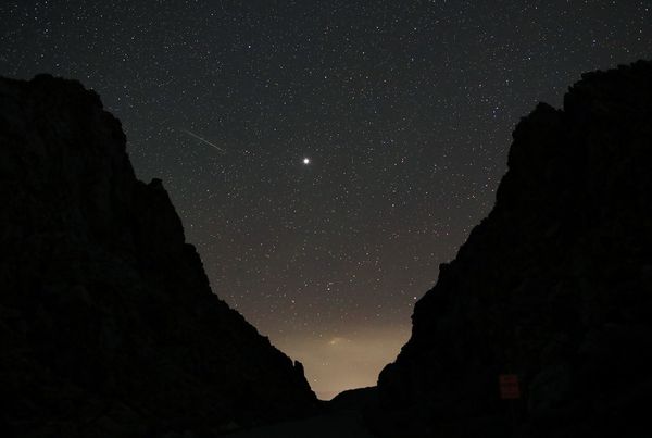 A long-exposure photograph of a starry night sky seen between the silhouetted walls of a canyon or mountains. The bright object near the center is Jupiter, and a meteor/shooting star streaks across the upper left sky.