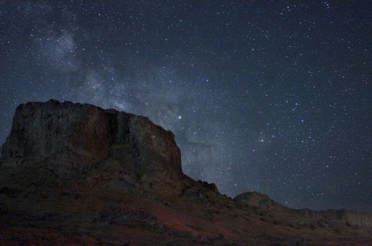 A wide-angle, low-light photograph captures a rugged mountain formation against a vast, star-filled night sky, with the Milky Way faintly visible. The mountain occupies the left half of the frame, its dark, textured face rising sharply. The right half of the image is dominated by the night sky, a deep blue-black adorned with countless pinpricks of light from stars.