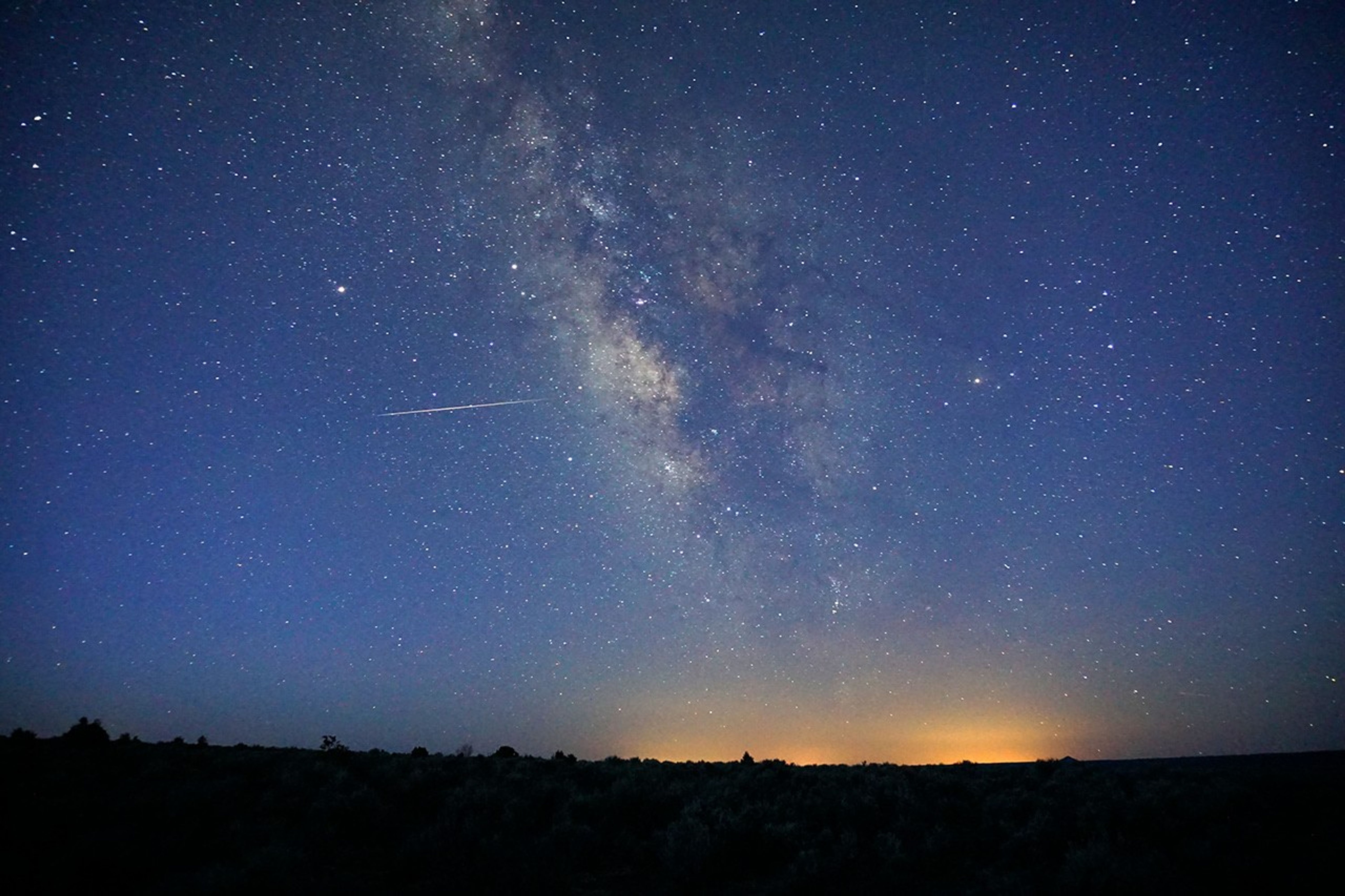 A bright meteor streaks across a star-filled night sky beside the Milky Way over a dark silhouetted landscape, illustrating the Lyrid meteor shower peaking April 21–22.