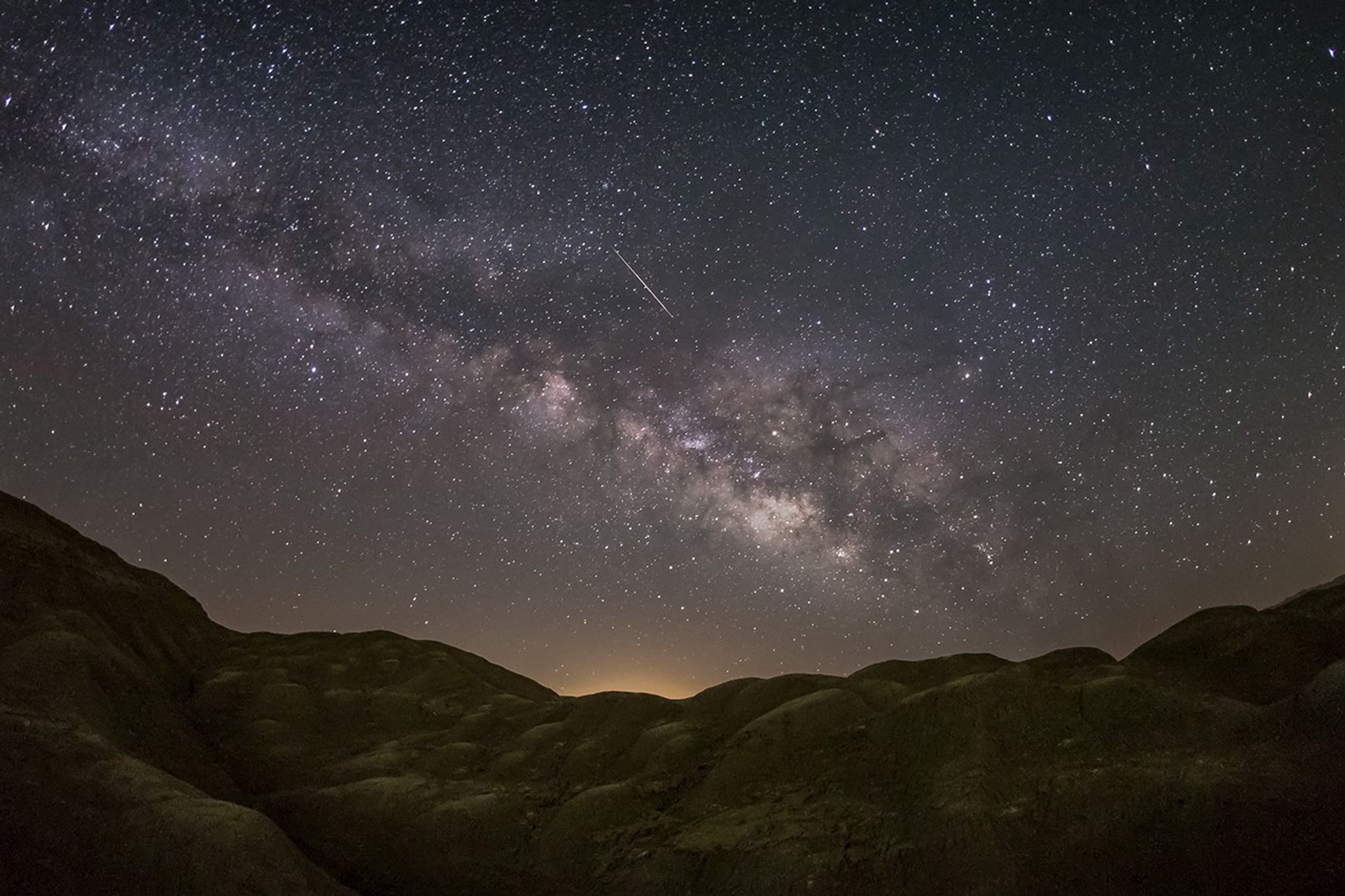 A meteor streaks near the Milky Way above dark desert hills, illustrating the Lyrid shower's peak viewing opportunity on the night of April 21st–22nd.