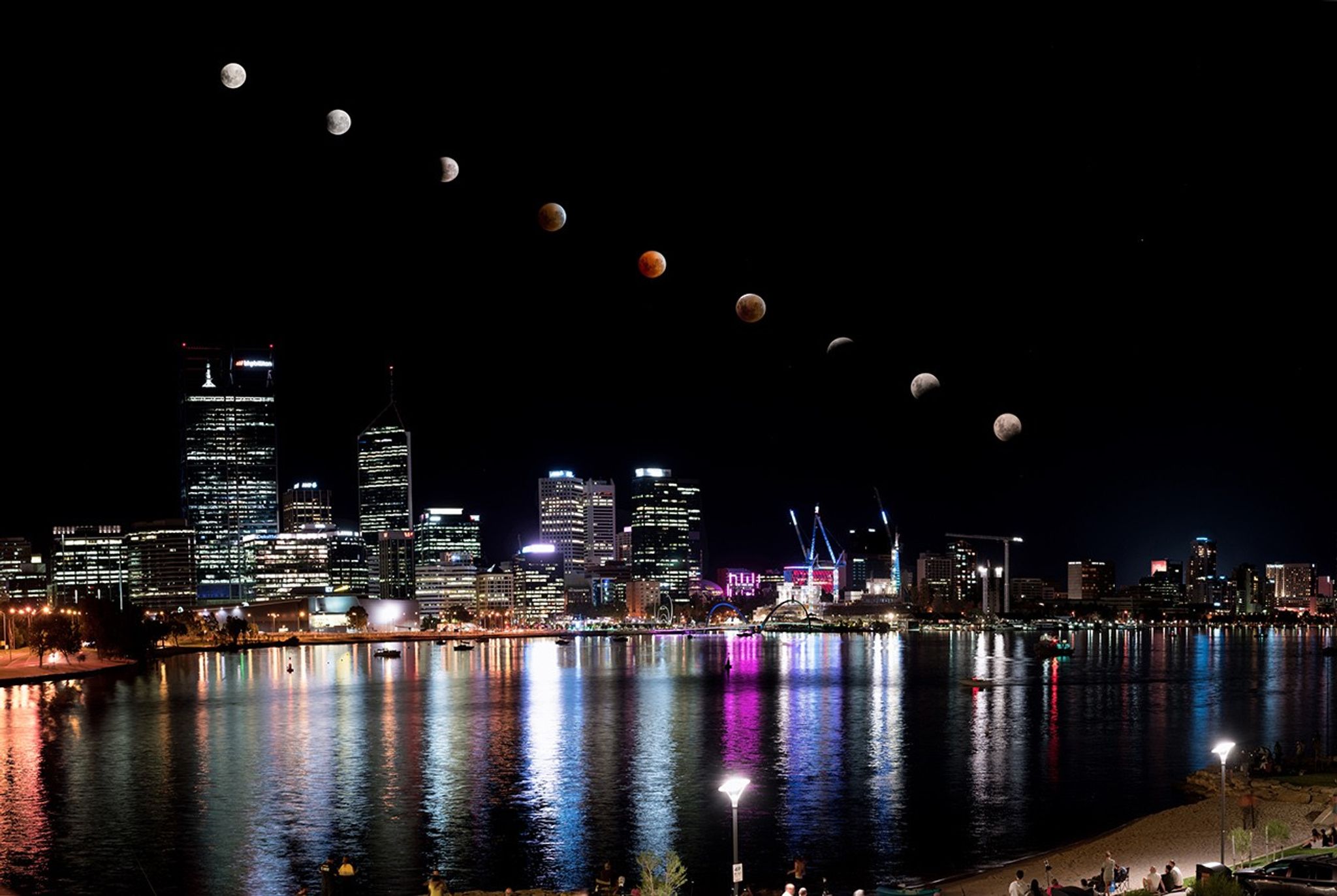 A wide-angle, night-time composite photograph of a lunar eclipse over the Perth, Australia skyline. A diagonal line of nine moons arches across the dark sky, documenting the progression of the eclipse: starting from a bright full moon at the top left, transitioning into a dark reddish-orange "Blood Moon" at the center, and returning to a partial moon at the bottom right. Below, the brightly lit city skyscrapers are reflected in the calm waters of the Swan River, with vibrant streaks of white, gold, and magenta light shimmering on the surface.