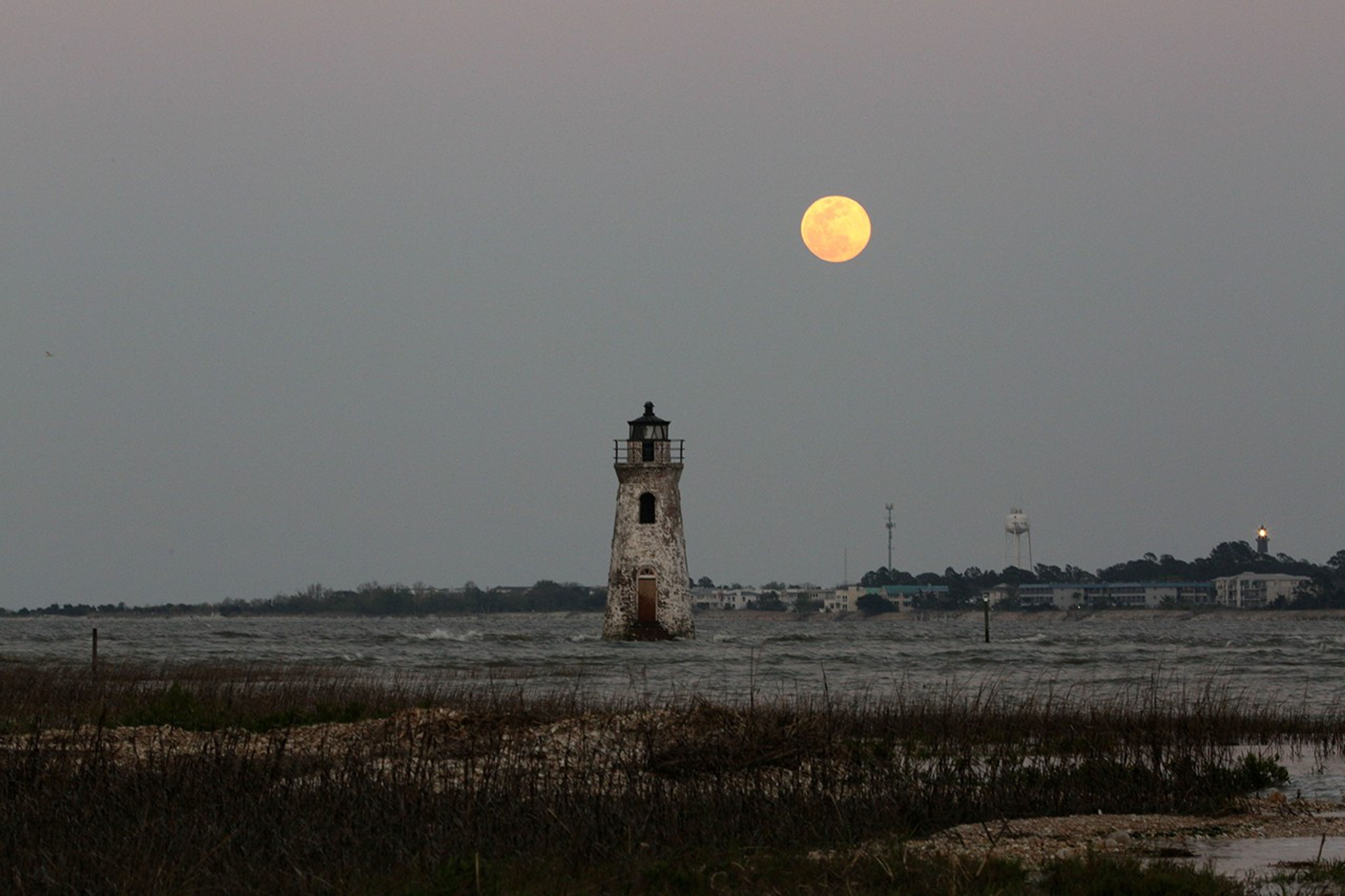 A red brick lighthouse with white patches over most of the brick sits in the water under a yellowish orange full moon. There are buildings and a water tower in the background. Tall grass is growing in the foreground.